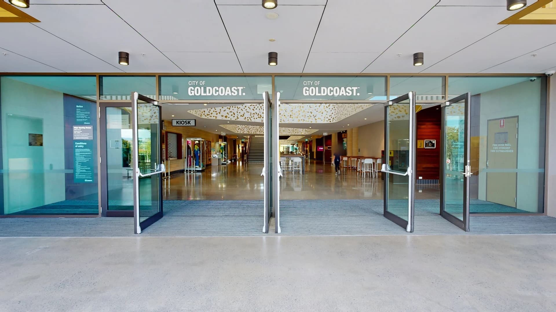 Glass doors open into a spacious interior with a polished floor. Above the entrance, "City of Gold Coast" is displayed. Inside, a kiosk is visible on the left, and tables with chairs are on the right. The ceiling features decorative lighting. In the background, a staircase leads to an upper level. The ceiling outside is white with recessed lighting, and the floor is light grey.