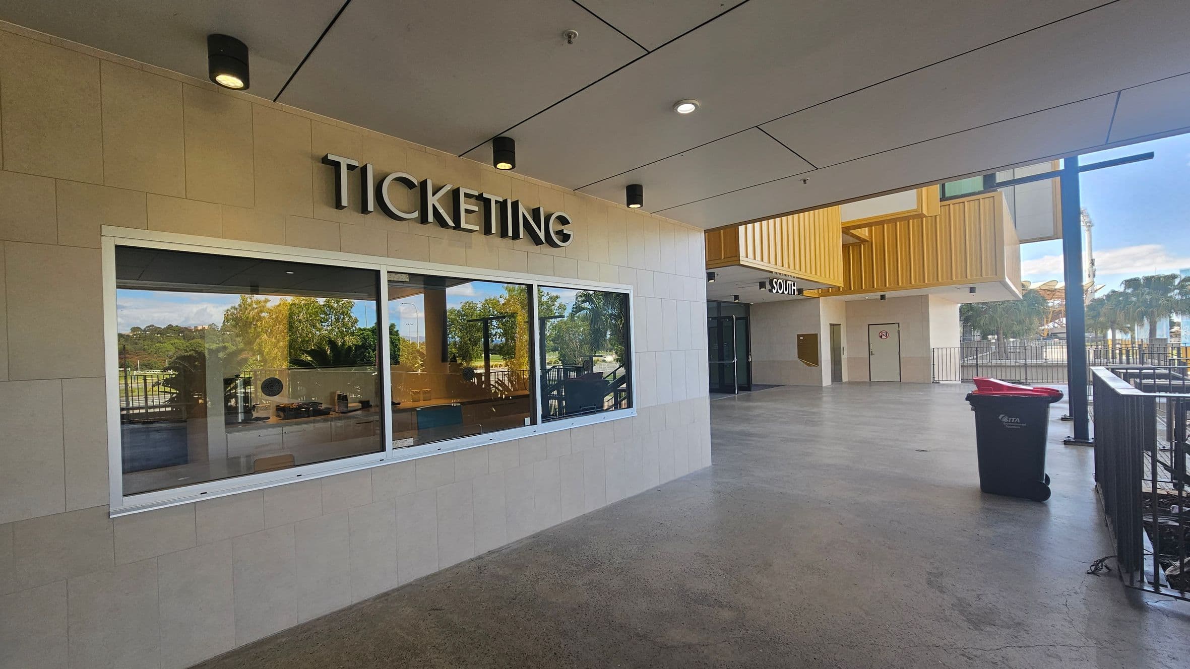 A ticketing area with a large window on the left, displaying the word "TICKETING" above it. The floor is grey and smooth. On the right, there is a corridor leading to a door marked "SOUTH" with a yellow structure above. A blue bin with a red lid is near the railing. The ceiling is light-coloured with recessed lights. In the background, there are trees and a glimpse of a Ferris wheel.