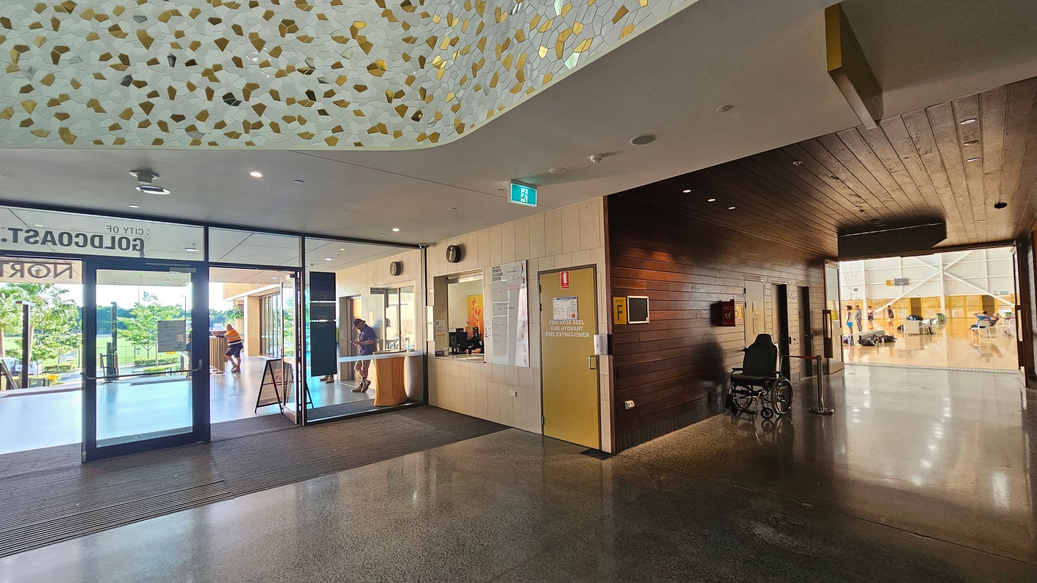 A modern indoor space with a polished concrete floor. On the left, large glass doors lead outside, showing greenery and a person in an orange shirt. In the centre, a reception area with a counter and a noticeboard. On the right, a hallway with wooden panelling leads to a bright room where people are seated at tables. The ceiling has a decorative pattern with gold and white shapes. A wheelchair is parked near the hallway entrance.