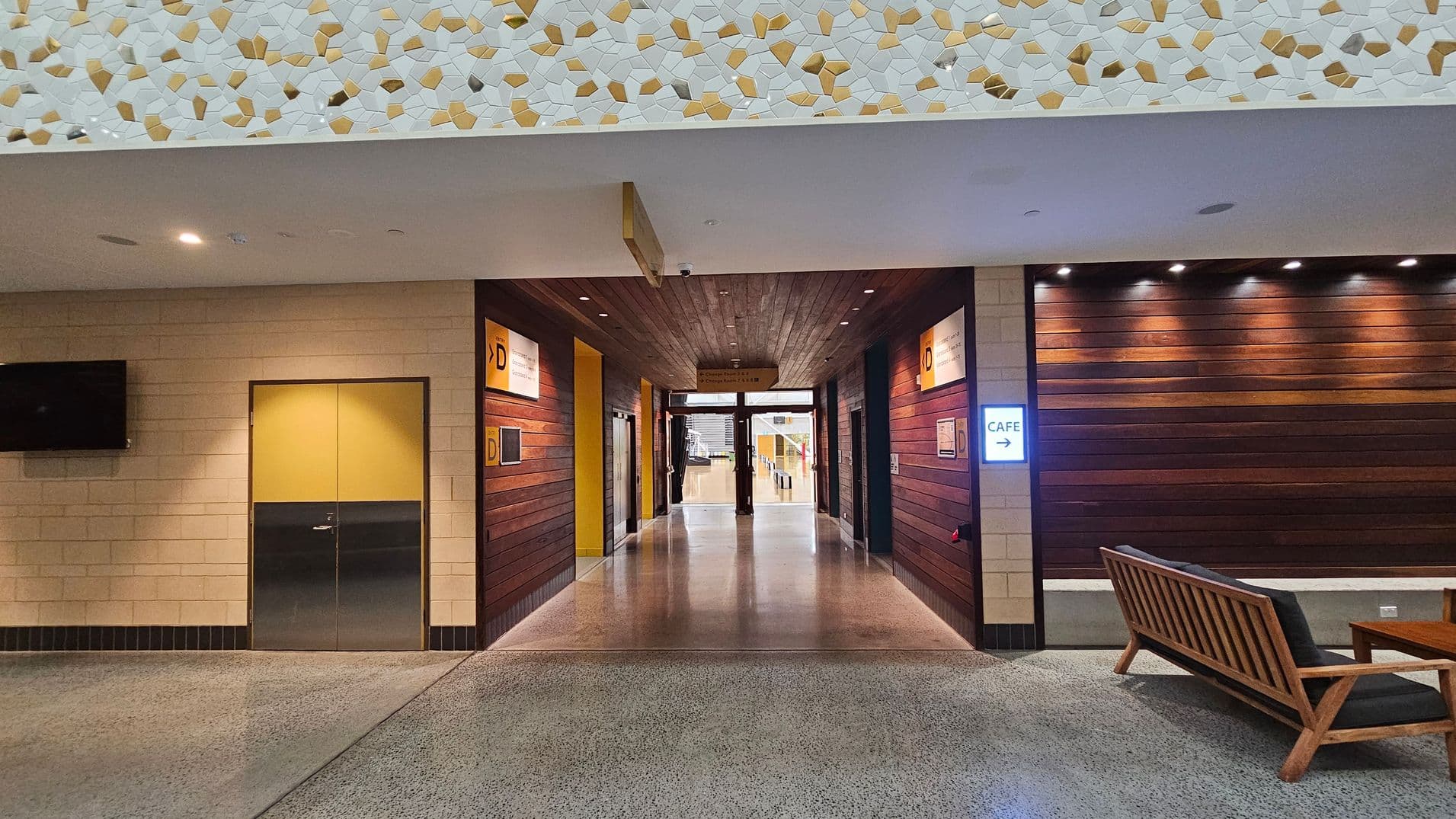 A hallway with a polished concrete floor and a wooden ceiling. On the left, there's a yellow and grey door next to a wall-mounted TV. On the right, a wooden bench with dark cushions is placed against a wall with horizontal wooden panels. A sign with "CAFE" and an arrow is illuminated. The upper section of the wall features a mosaic pattern with white and gold tiles. The hallway leads to a glass door at the far end.