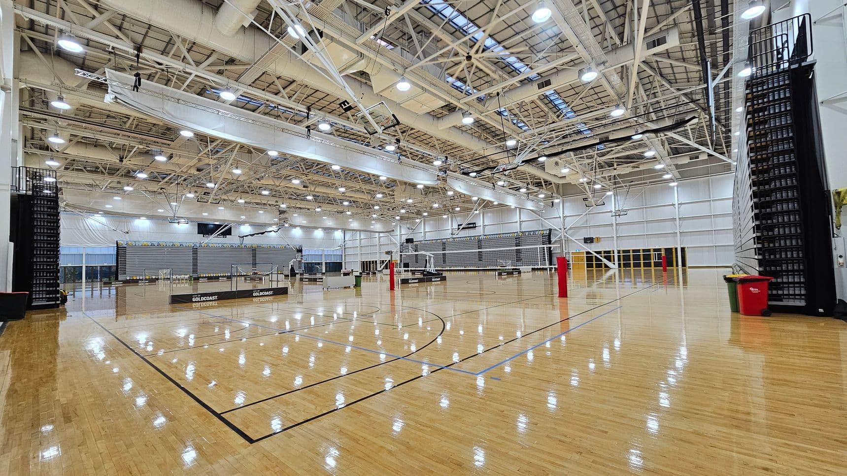 A spacious indoor sports hall with a polished wooden floor and high ceiling. Volleyball nets are set up across the court, with red poles supporting them. On the left and right, there are retractable bleachers. The ceiling features exposed beams and numerous lights. In the background, large windows and doors allow natural light to enter. A red bin is visible on the right side near the bleachers.