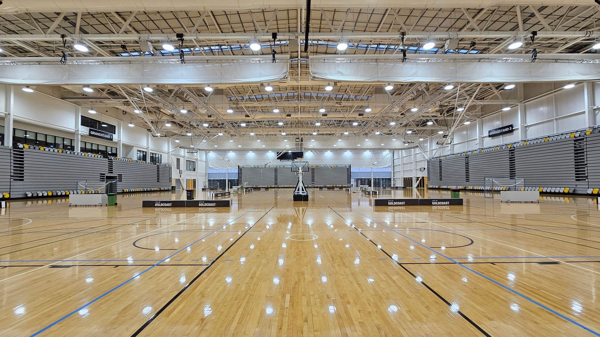 A spacious indoor sports hall with a polished wooden floor marked for basketball. In the centre, a basketball hoop stands prominently. On both sides, there are large, empty grandstands with grey seating. The ceiling is high with exposed beams and numerous lights. The walls are lined with windows and banners that read "GOLDCOAST." The overall atmosphere is bright and open.