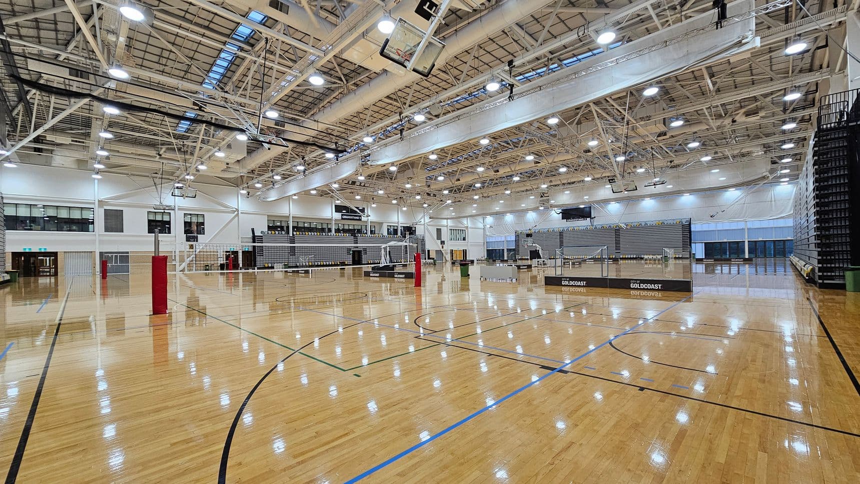 A large indoor sports hall with a polished wooden floor marked for various sports. Volleyball nets are set up across the court. The ceiling is high with exposed beams and numerous lights. On the left, there are windows and doors, while on the right, there are retractable bleachers. In the background, more seating and a scoreboard are visible. The space is well-lit and spacious, suitable for multiple sports activities.