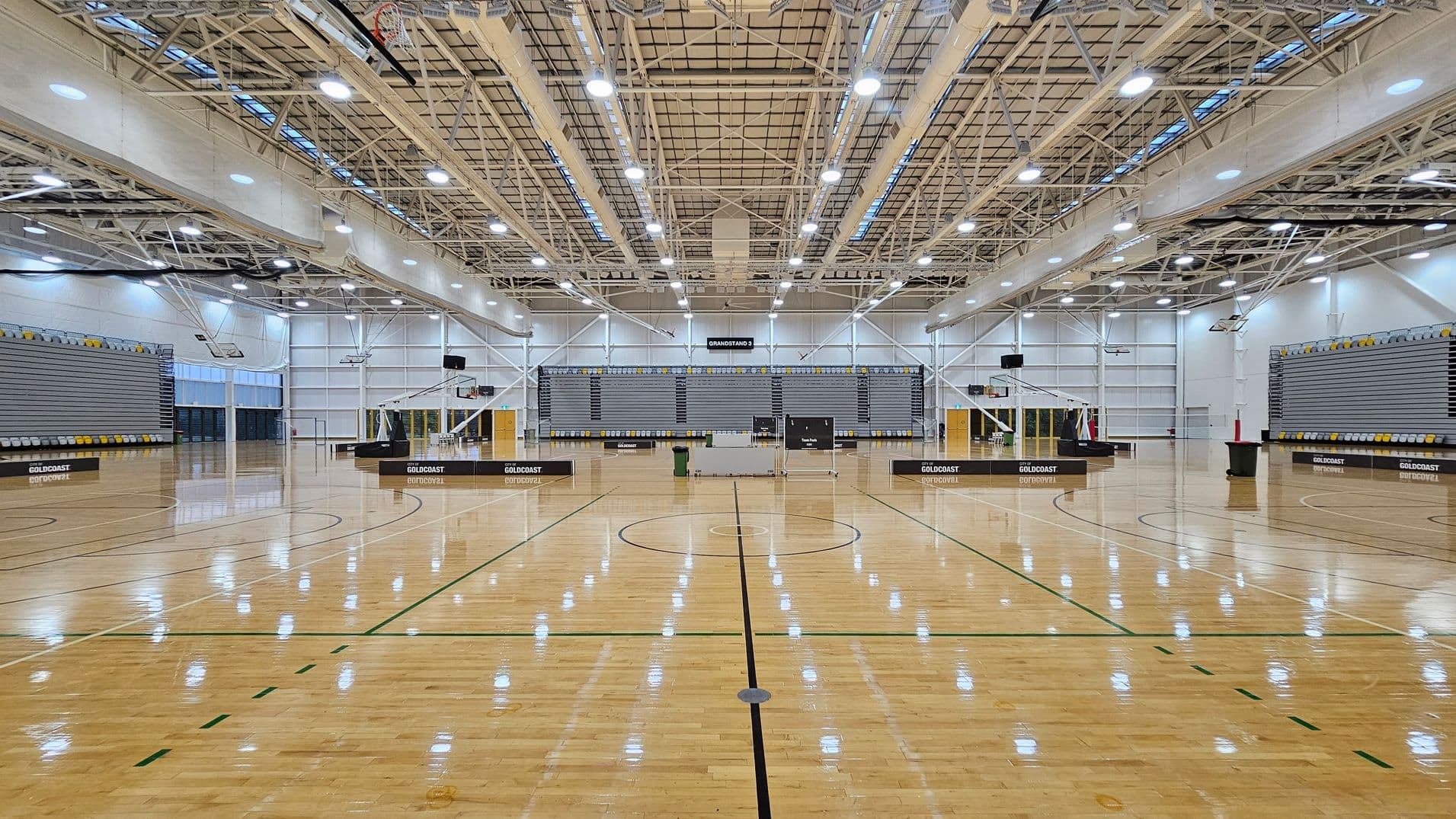 A large indoor sports hall with a polished wooden floor marked for basketball. In the centre, there are two basketball hoops. The background features a grandstand with grey and yellow seating. The ceiling is high with exposed beams and numerous lights. The walls are white with large windows on the left side, allowing natural light. The word "GOLDCOAST" is visible on the equipment and seating areas.