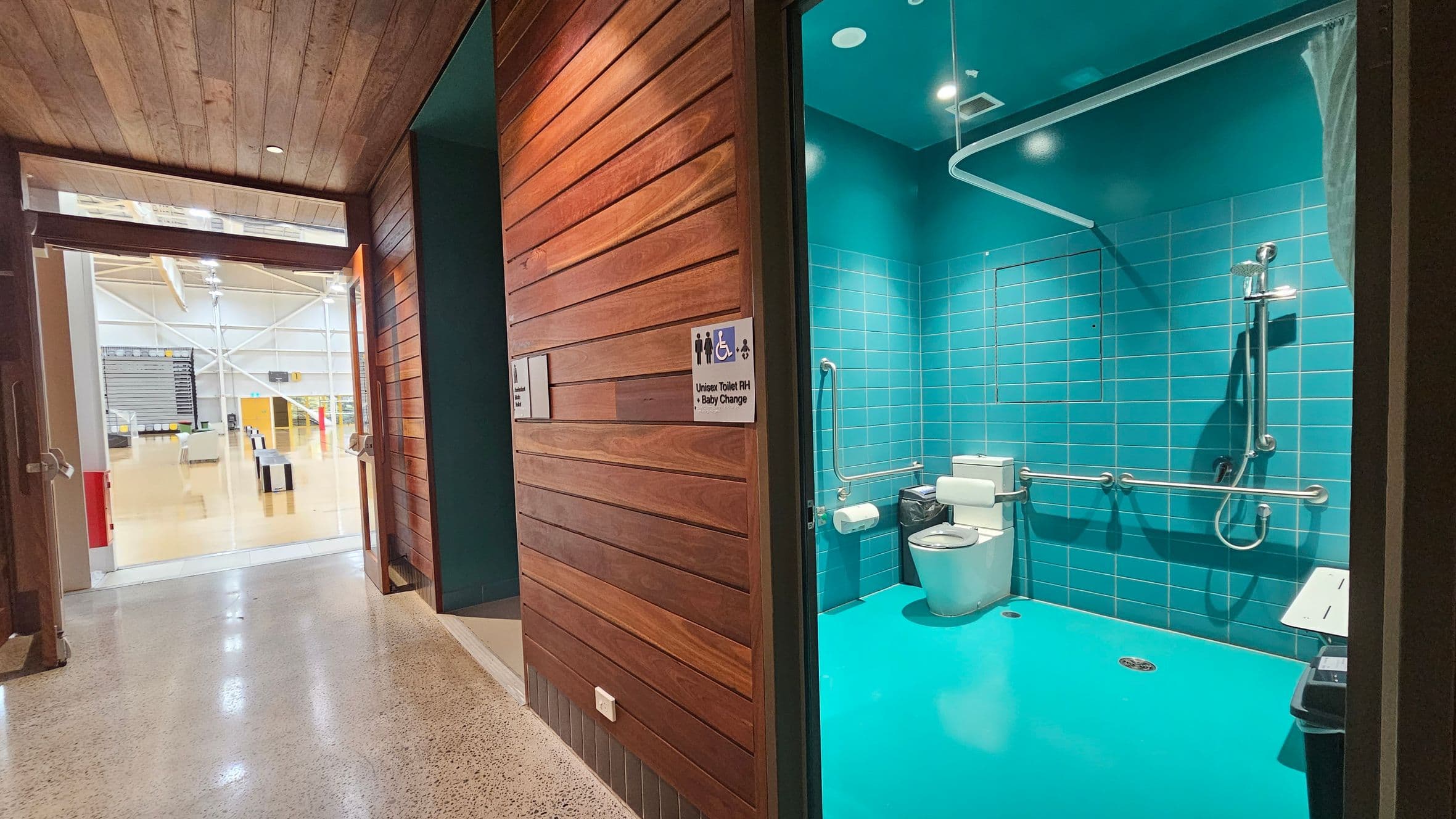 A modern, accessible unisex toilet with baby change facilities is on the right, featuring teal tiles and a bright teal floor. It includes a toilet, grab bars, a shower, and a baby changing station. The entrance has wooden panelling and a sign indicating the facilities. On the left, through the open door, is a spacious area with a polished concrete floor, visible seating, and a high ceiling with exposed beams and lights.
