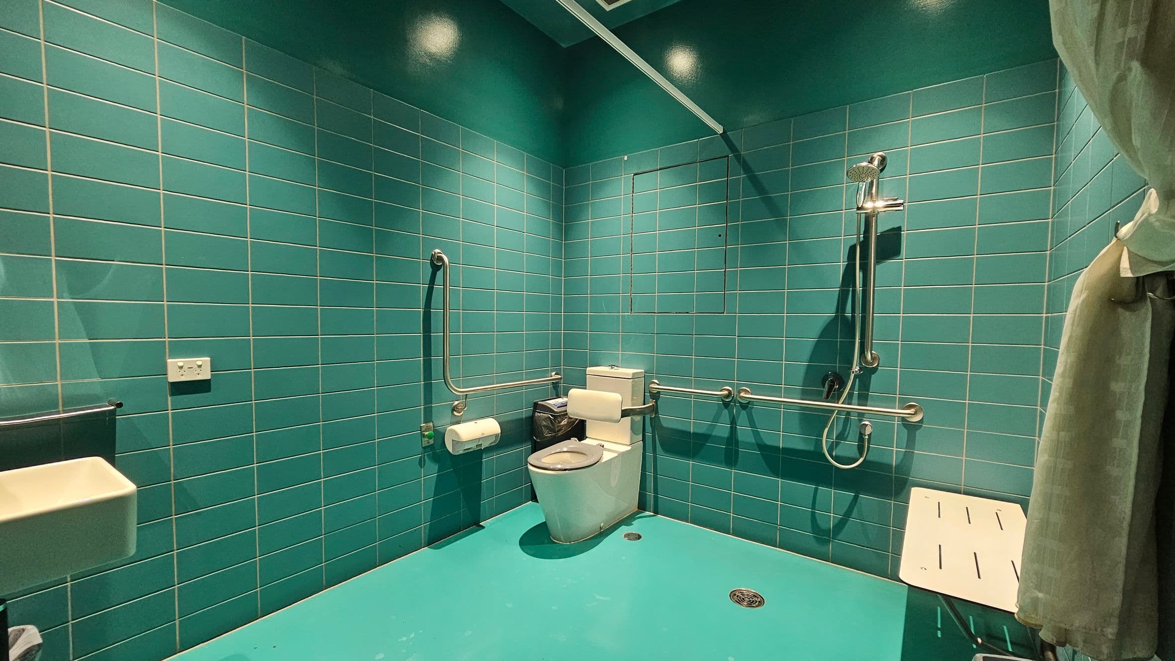 A bathroom with teal tiles on the walls and a matching teal floor. In the centre, there is a toilet with grab bars on both sides. To the right, a shower area with a handheld showerhead and a white foldable shower seat. A curtain is partially visible on the far right. On the left, a small white sink and a power outlet are present. The ceiling is also teal, with a light fixture above.