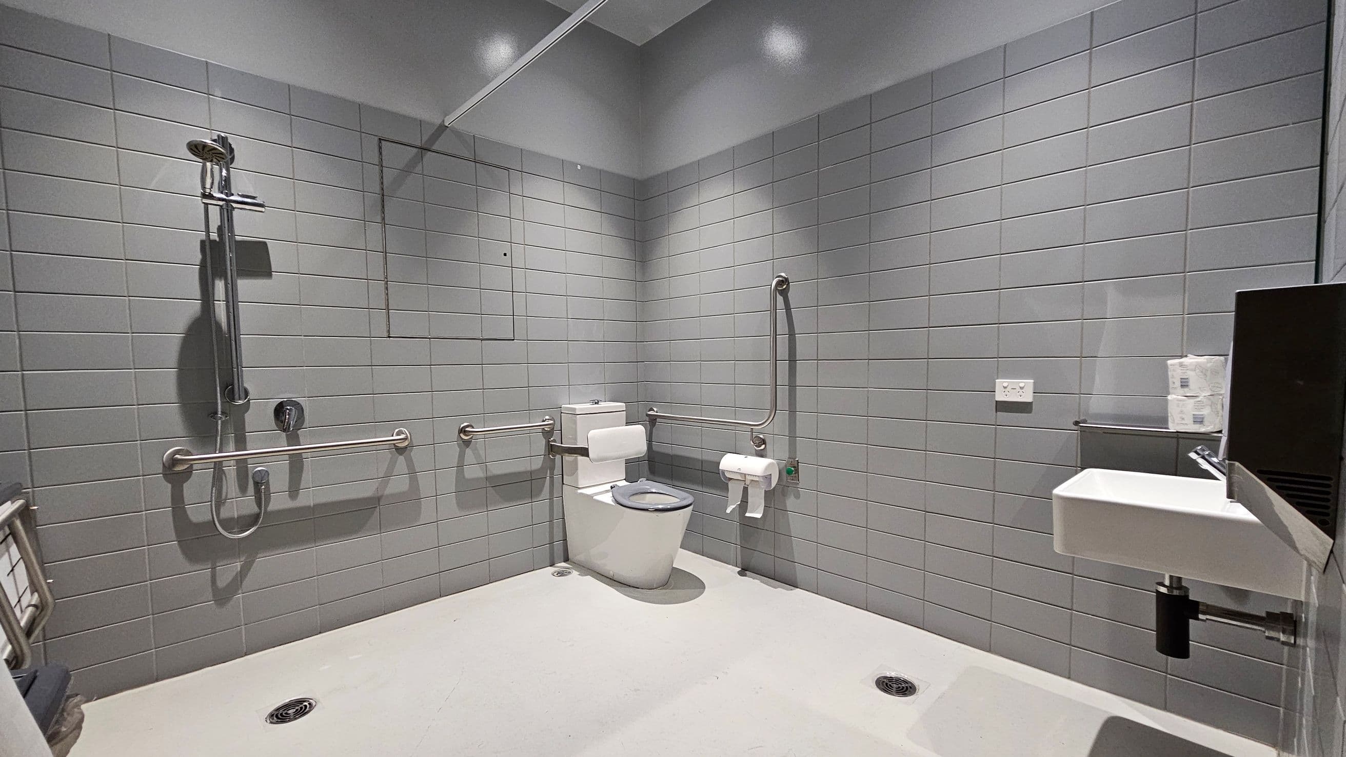 A spacious, accessible bathroom with grey tiled walls and a white floor. On the left, there is a shower with a handheld showerhead and a horizontal grab bar. In the centre, a toilet is equipped with multiple grab bars and a toilet paper holder. On the right, a small white sink is mounted on the wall with a soap dispenser above it. There is a power outlet and a shelf holding toilet paper rolls nearby. The ceiling is plain with recessed lighting.