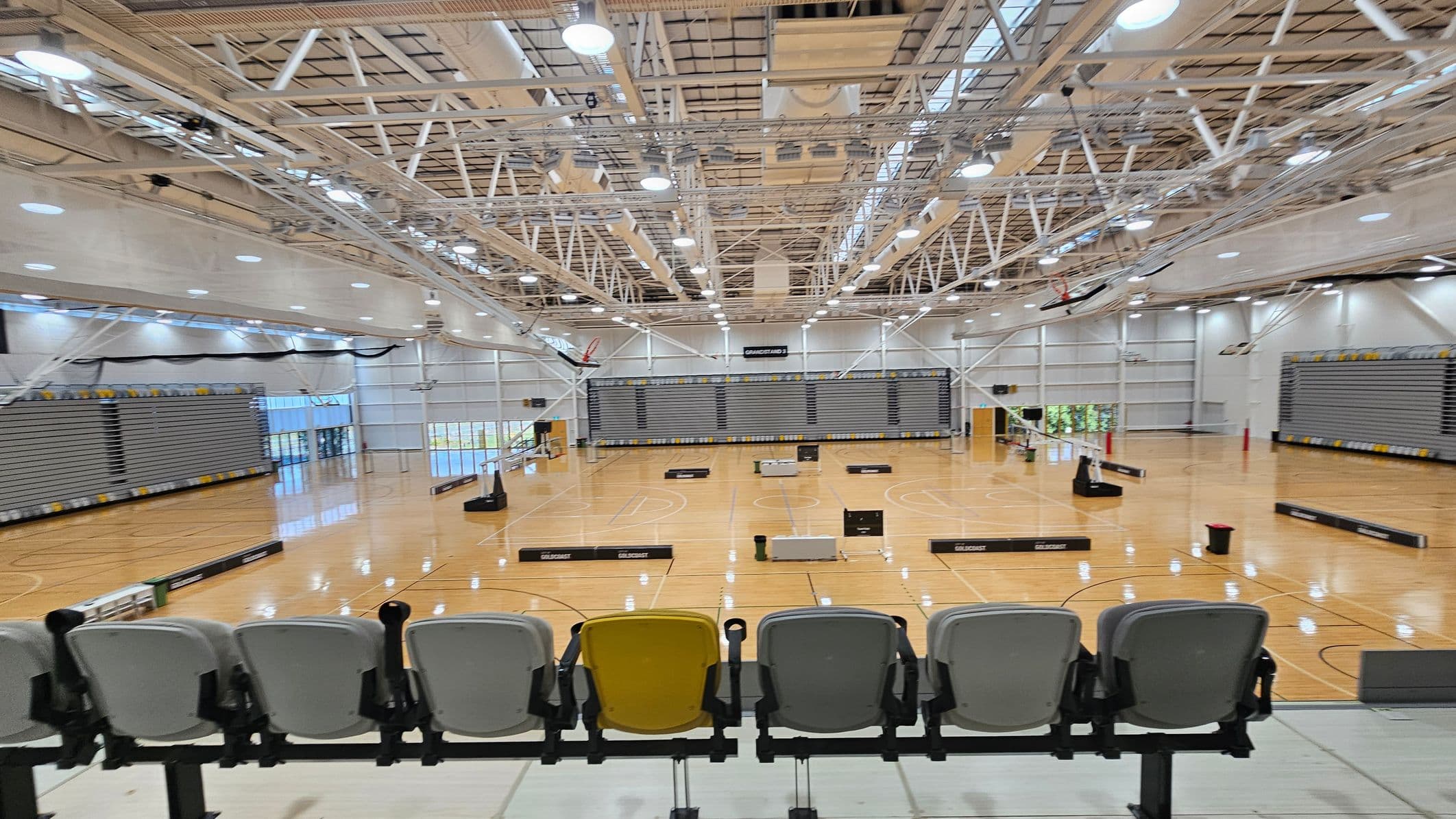 A large indoor sports hall with a polished wooden floor and multiple basketball hoops. The ceiling is high with exposed beams and numerous lights. In the foreground, there are rows of grey and yellow seats. The background features retractable bleachers along the walls. Various sports equipment is scattered across the floor, and large windows on the left allow natural light to enter.