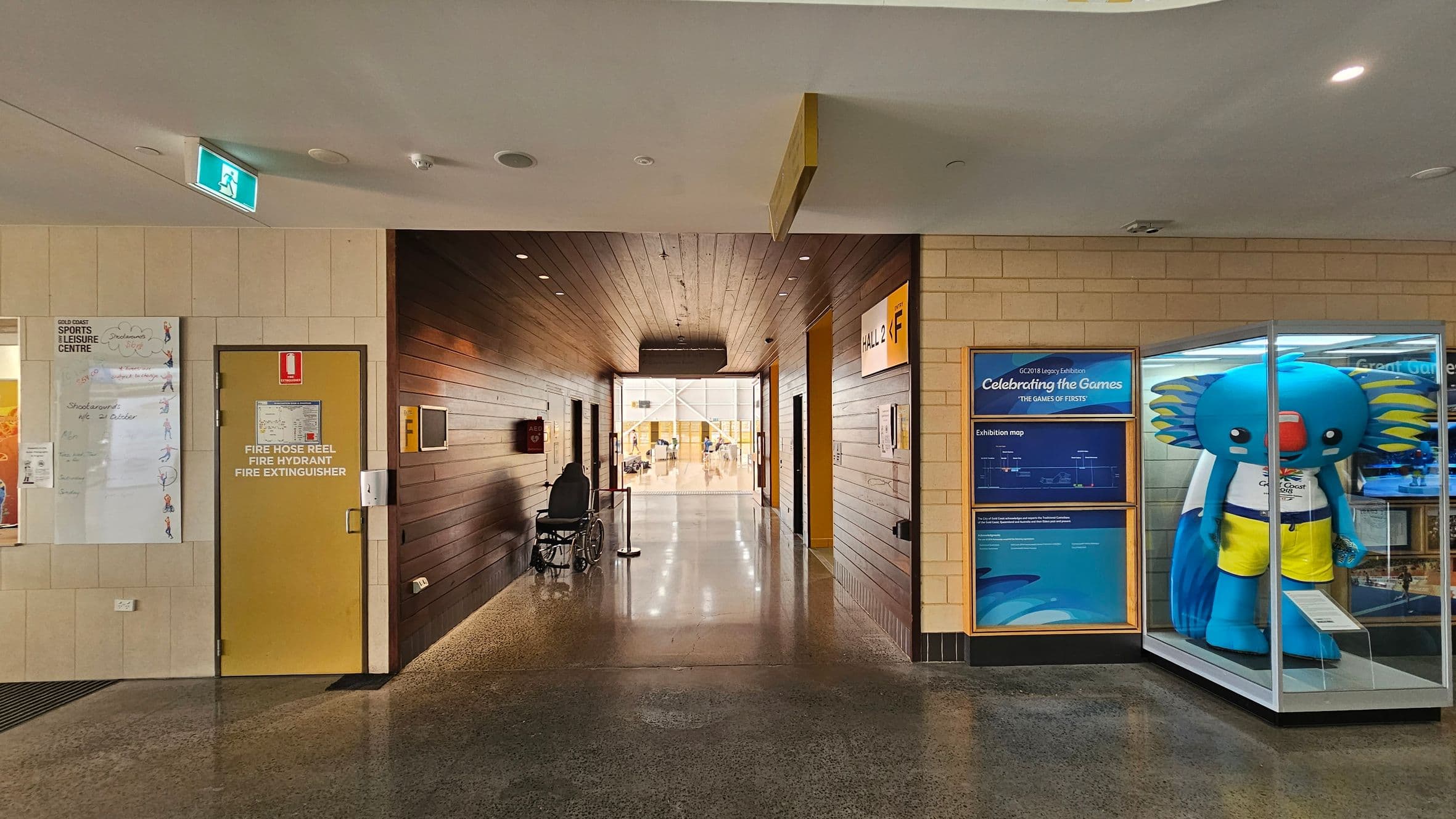 A hallway with a polished concrete floor and a wooden ceiling. On the left, there's a yellow door marked for fire safety equipment, with a notice board nearby. A wheelchair is positioned against the left wall. On the right, a large blue mascot figure is displayed in a glass case, with exhibition information about the Gold Coast Games. The corridor leads to a bright area with people visible in the distance.