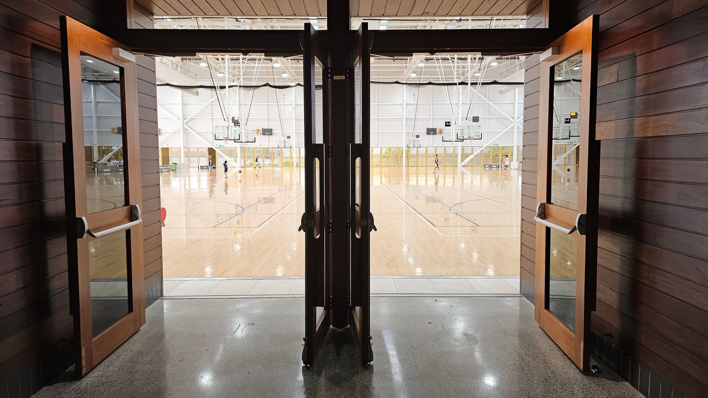 Wooden double doors with glass panels are open, leading into a large indoor sports hall. The floor is polished wood, and several basketball hoops are visible. People are playing basketball in the distance. The ceiling is high with exposed beams and lighting fixtures. The foreground features a polished concrete floor.