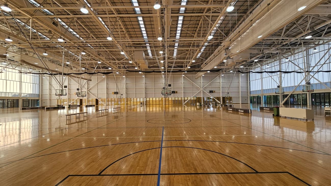 A large indoor sports hall with a polished wooden floor marked for basketball. Multiple basketball hoops are suspended from the ceiling, which is high and features exposed beams and lighting. The walls are lined with tall windows, allowing natural light to flood the space. On the left and right, there are entrances and seating areas. The background shows a series of doors and partitions.