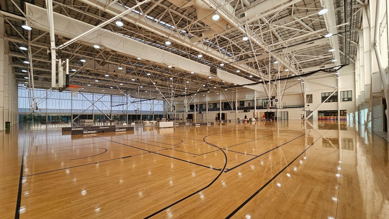 A spacious indoor basketball court with a polished wooden floor and black court markings. On the left, there are basketball hoops and a large windowed wall. The ceiling is high with exposed beams and numerous lights. On the right, there are more hoops and a few people in the distance. The background features a mezzanine level with windows and doors. The overall space is well-lit and expansive.