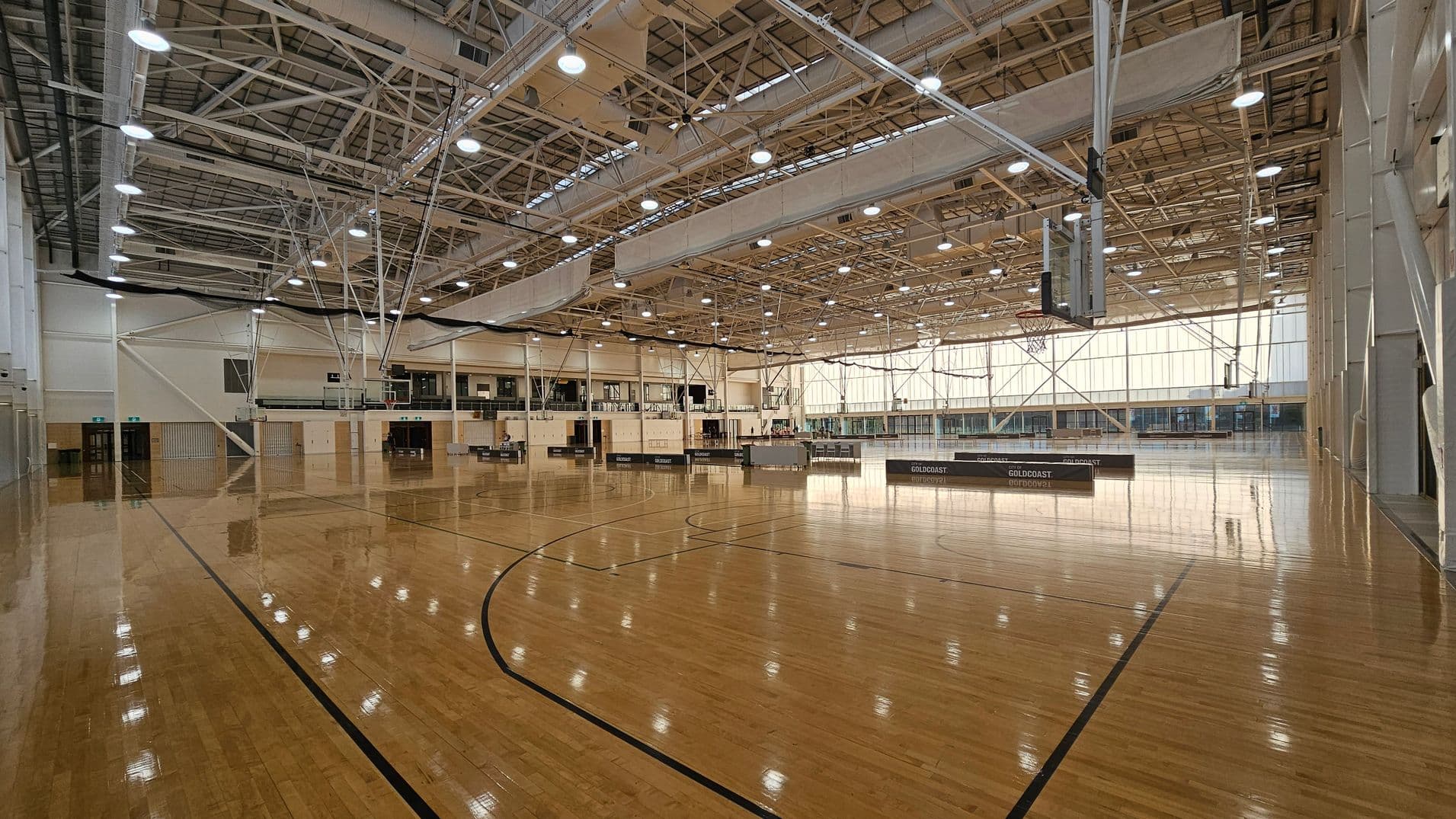 A large indoor basketball court with a polished wooden floor. On the right, a basketball hoop is visible. The ceiling is high with exposed beams and numerous lights. The left side has doors and walls with light-coloured panels. In the background, there are more basketball hoops and a series of windows allowing natural light to enter. The space is open and expansive, suitable for multiple sports activities.