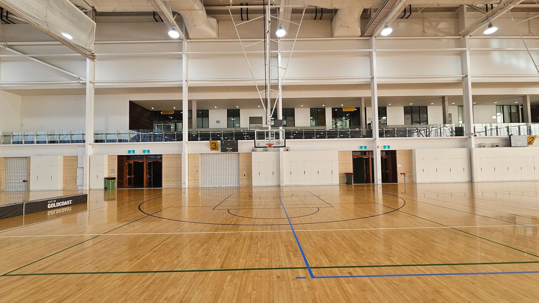 A spacious indoor basketball court with a polished wooden floor and marked lines. In the centre is a basketball hoop with a backboard. The background features a two-storey structure with large windows and a balcony railing. There are multiple doors on the ground level, and a sign on the left reads "City of Gold Coast." The ceiling is high with exposed beams and lighting fixtures.