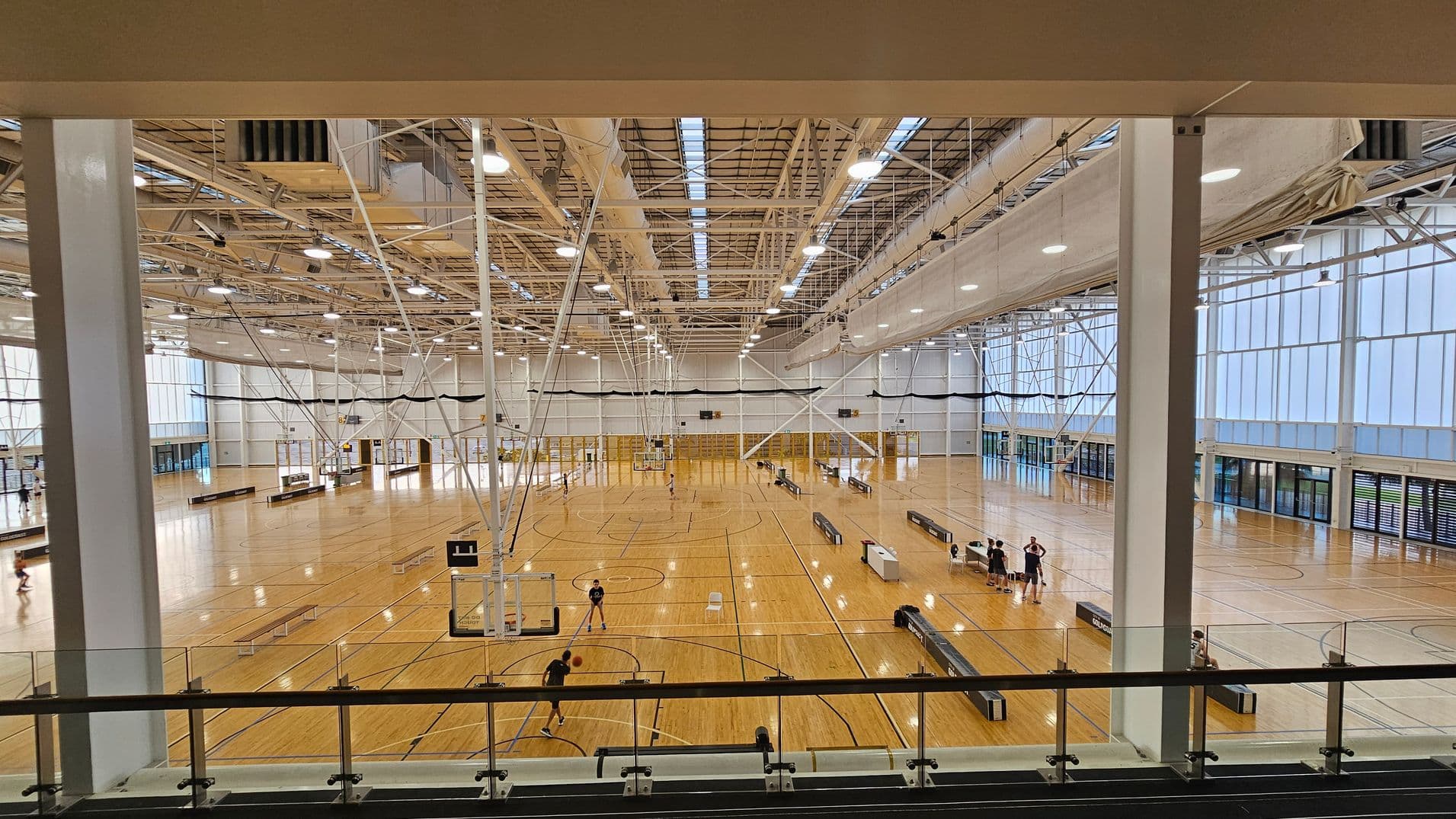 A large indoor sports hall with a wooden floor and multiple basketball courts. The ceiling is high with exposed beams and lighting fixtures. On the left, a few people are playing basketball. In the centre, there are more courts with scattered players. On the right, a group of people is gathered near a court. The walls are lined with large windows, allowing natural light to fill the space. A glass railing is visible in the foreground.