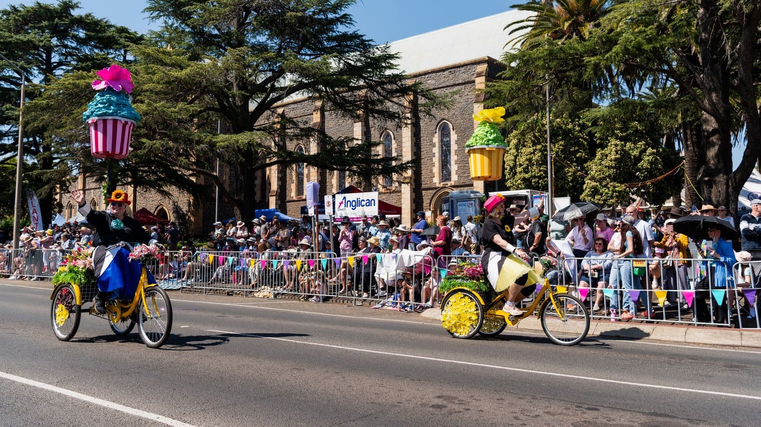 On the left, a person rides a yellow tricycle adorned with flowers and a large cupcake decoration. On the right, another person on a similar tricycle with a different cupcake decoration. Both are on a road lined with colourful bunting. In the background, a crowd stands behind metal barriers, some holding umbrellas. Behind them is a stone building with arched windows, partially obscured by large trees. The scene is lively and festive.