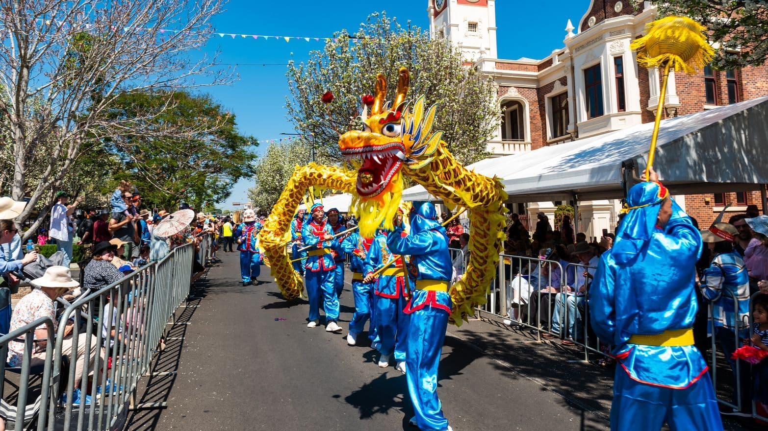 A vibrant street parade with performers in blue costumes manipulating a large, colourful dragon puppet. The dragon is yellow with red and blue accents. On the left, spectators sit and stand behind metal barriers, some wearing hats. On the right, a white tent is set up in front of a historic brick building with a clock tower. Trees line the street, and the sky is clear and blue. Bunting is strung across the street above the parade.