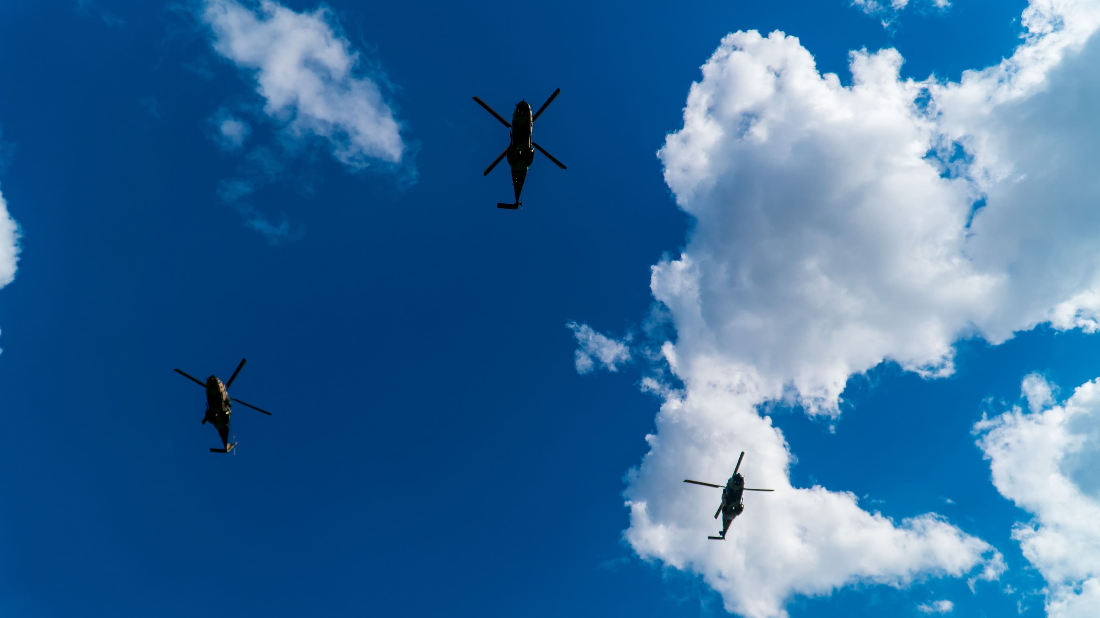 Three helicopters are flying in formation against a bright blue sky. One helicopter is positioned in the centre, with the other two on the left and right. White clouds are scattered across the sky, with larger clouds on the right side. The helicopters are silhouetted, making their rotors and tails clearly visible.