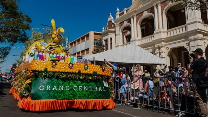 A colourful parade float adorned with vibrant yellow and orange flowers and a large floral arrangement on top, labelled "Grand Central," moves along a street. On the right, a crowd of spectators stands behind metal barriers, some taking photos. Behind them is a white tent and a historic building with ornate architecture. The sky is clear and blue, and the street is lined with trees.