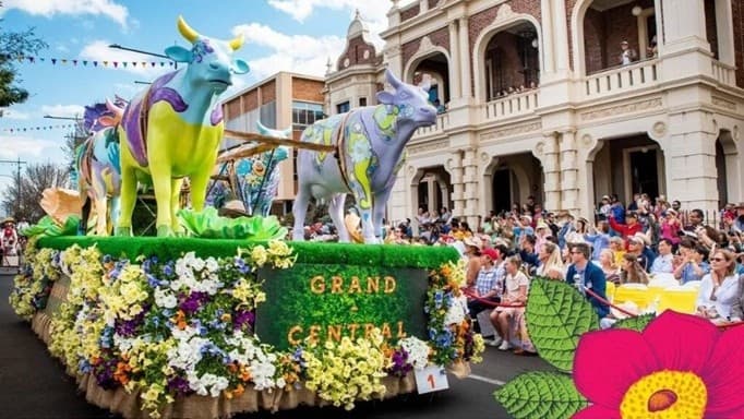 A vibrant parade float adorned with large, colourful cow sculptures and lush greenery moves along a street. The float is decorated with an abundance of flowers in various colours, and the words "GRAND CENTRAL" are visible. On the right, a crowd of spectators sits on the pavement in front of a grand, historic building with arches and balconies. The street is lined with festive bunting overhead.