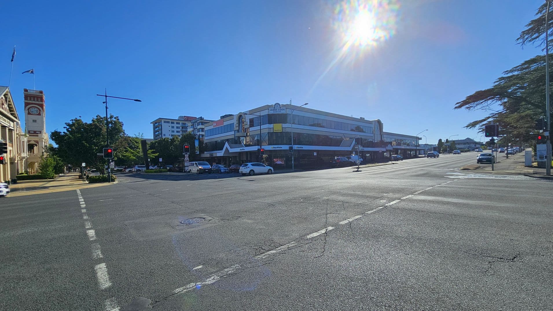A wide street intersection with traffic lights on each corner. On the left, there's a historic building with a clock tower and flags. Trees line the pavement. In the centre, a modern, multi-storey building with signage and parked cars. On the right, more trees and vehicles are visible. The road surface is marked with white lines and a manhole cover. The sun is bright, casting shadows across the scene.
