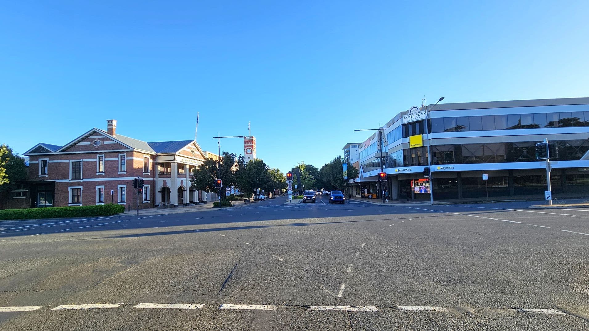 A street intersection with a historic brick building on the left, featuring a clock tower and arched entrance. On the right, a modern glass-fronted building with signage for "Ray White" and "James Cook." Traffic lights are visible at the intersection, and a few cars are on the road. Trees line the street in the background. The road surface is asphalt with visible lane markings.