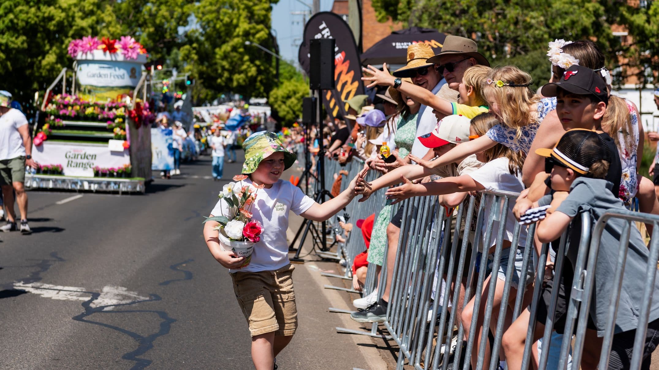 A parade scene with a child in the foreground wearing a floral hat and holding a pot of flowers, reaching out to high-five spectators behind a metal barrier on the right. The crowd includes adults and children, some wearing hats. In the background, a colourful float decorated with flowers and a sign reading "Garden Range" is visible. The street is lined with trees, and the atmosphere is lively and festive.