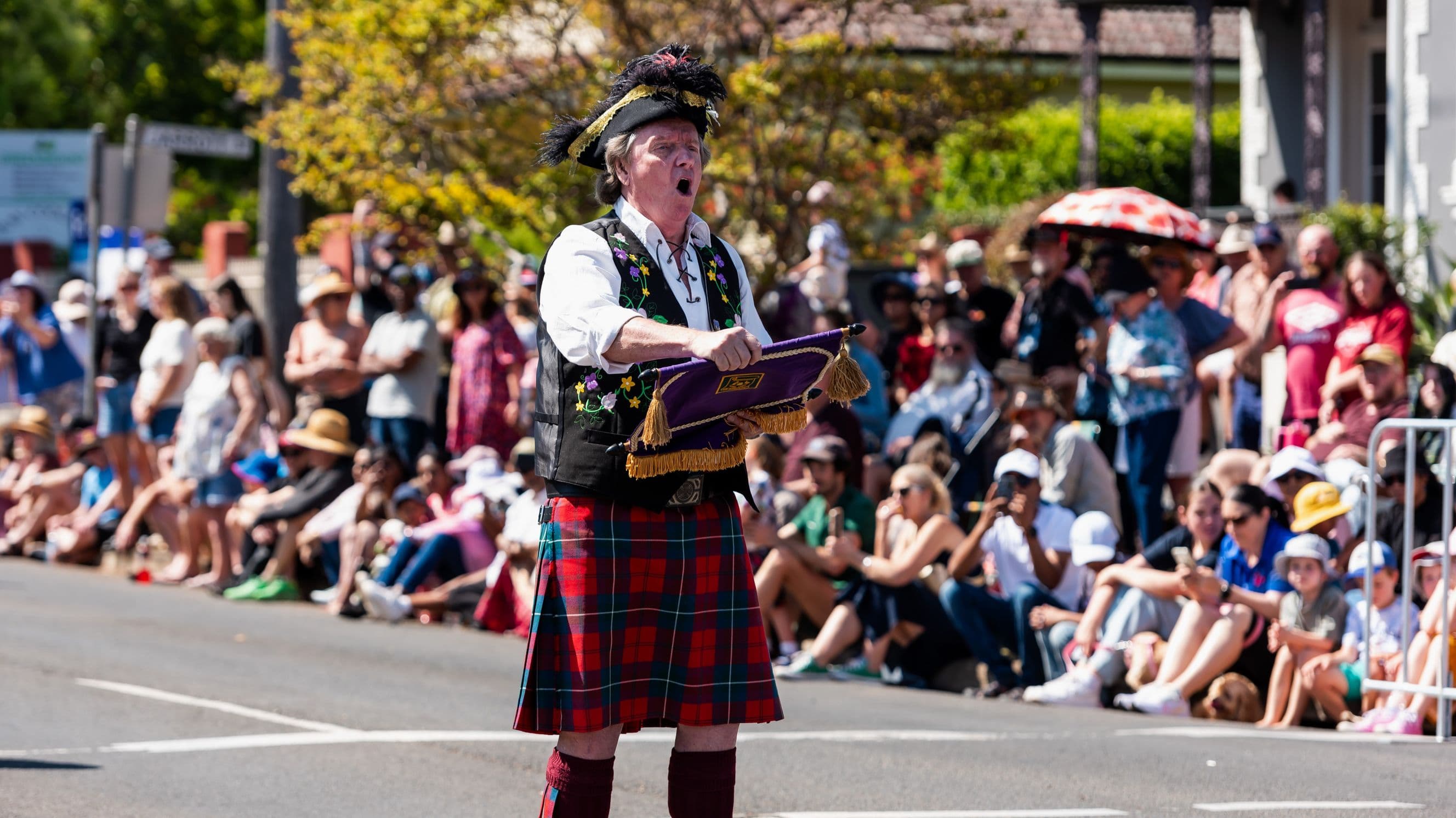 A person dressed in traditional Scottish attire, including a tartan kilt and a feathered hat, stands in the foreground holding a purple cushion with tassels. They appear to be speaking or singing. In the background, a large crowd of people, some seated and others standing, watch the event. Many are wearing hats and casual clothing, and some hold umbrellas for shade. The scene takes place on a street with trees and buildings visible in the background.