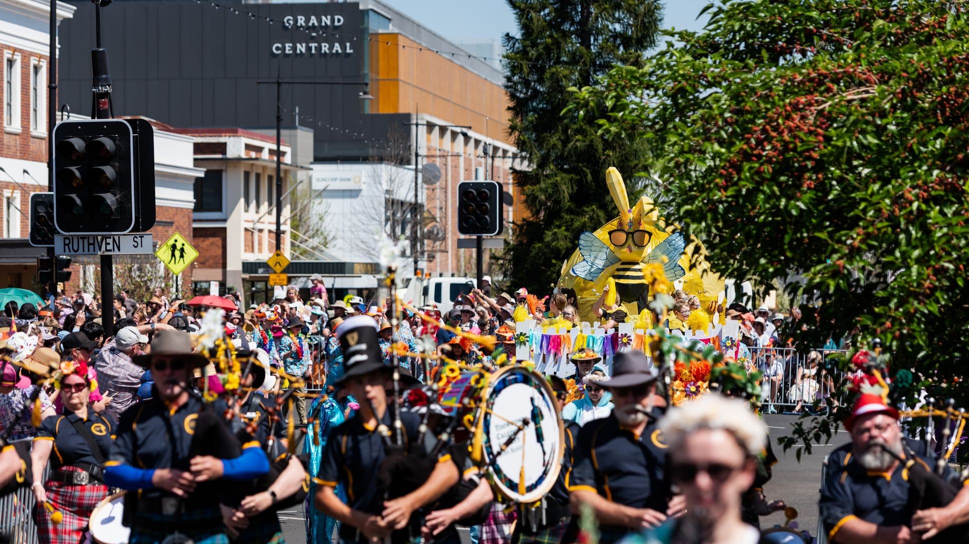 A lively street parade with a marching band in the foreground, featuring musicians in dark uniforms and hats. Behind them, a large float with a giant yellow bee wearing sunglasses is surrounded by people in colourful attire. The background shows a building with "GRAND CENTRAL" signage and a street sign for "RUTHVEN ST". Trees with green foliage are visible on the right, and a crowd lines the street, some holding umbrellas for shade.
