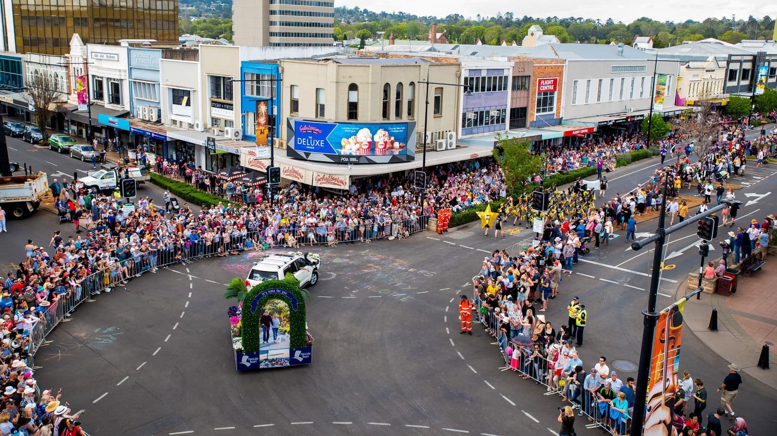 A bustling street parade with a large crowd gathered on both sides of the road, separated by metal barriers. In the centre, a white vehicle with a decorative arch on its roof moves along the street. People are dressed in casual attire, some wearing hats. Buildings with various shopfronts and advertisements line the background, including a prominent ice cream billboard. The road is marked with dashed lines, and there are traffic lights and street signs visible.