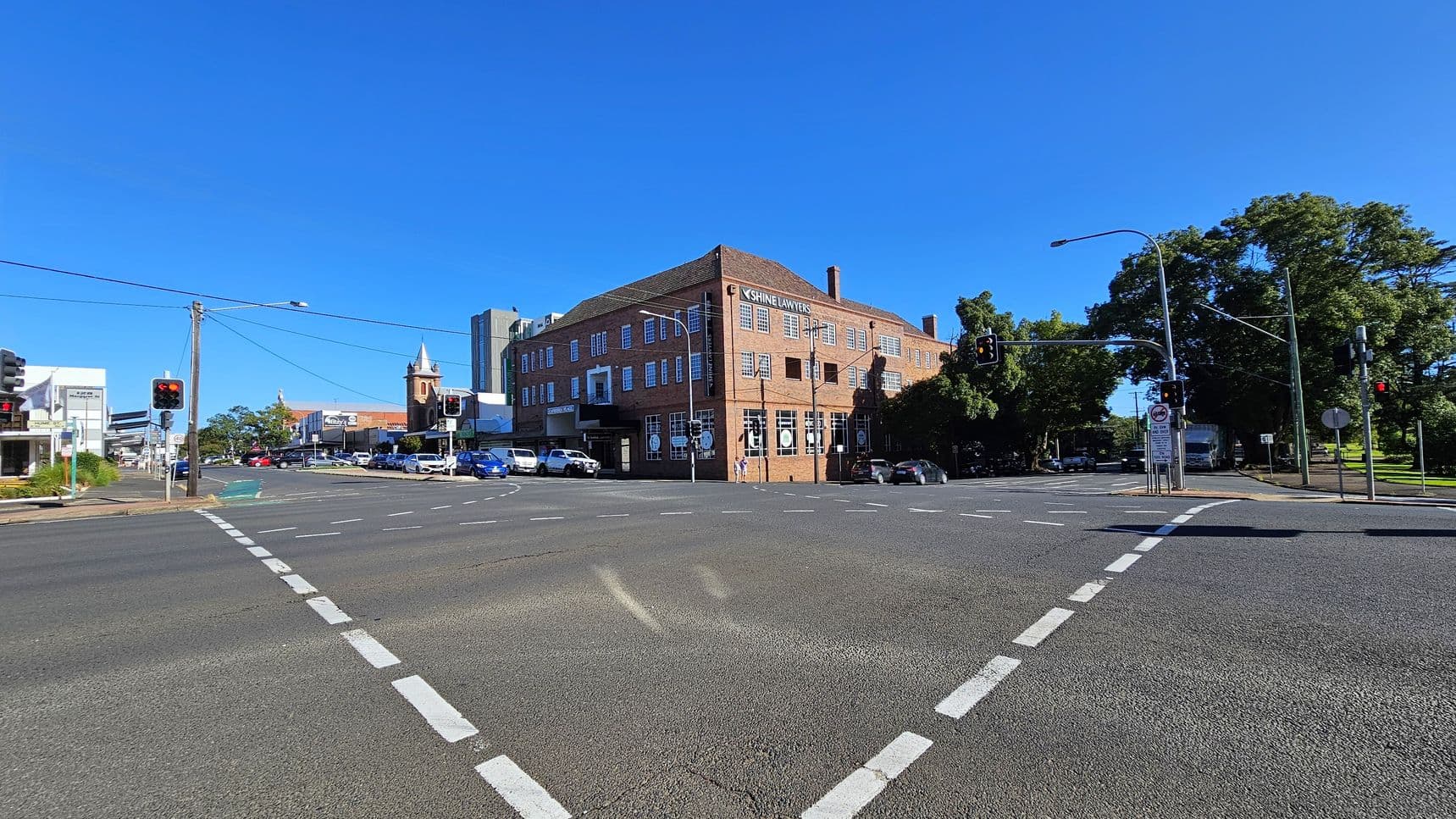 A street intersection with traffic lights and pedestrian crossings. On the right, a large brick building with "Shine Lawyers" signage. To the left, a church with a pointed tower and several modern buildings. Cars are parked along the street, and trees are visible in the background. The road surface is marked with white dashed lines, and the sky is clear.