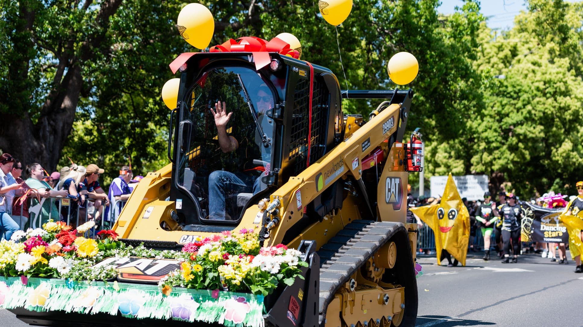 A parade scene with a yellow construction vehicle adorned with colourful flowers and yellow balloons. A person inside waves through the cab window. On the left, spectators stand behind a barrier, watching the parade. In the background, a person in a large star costume with a smiley face is visible, along with others holding a banner. Tall green trees line the street, providing a lush backdrop.