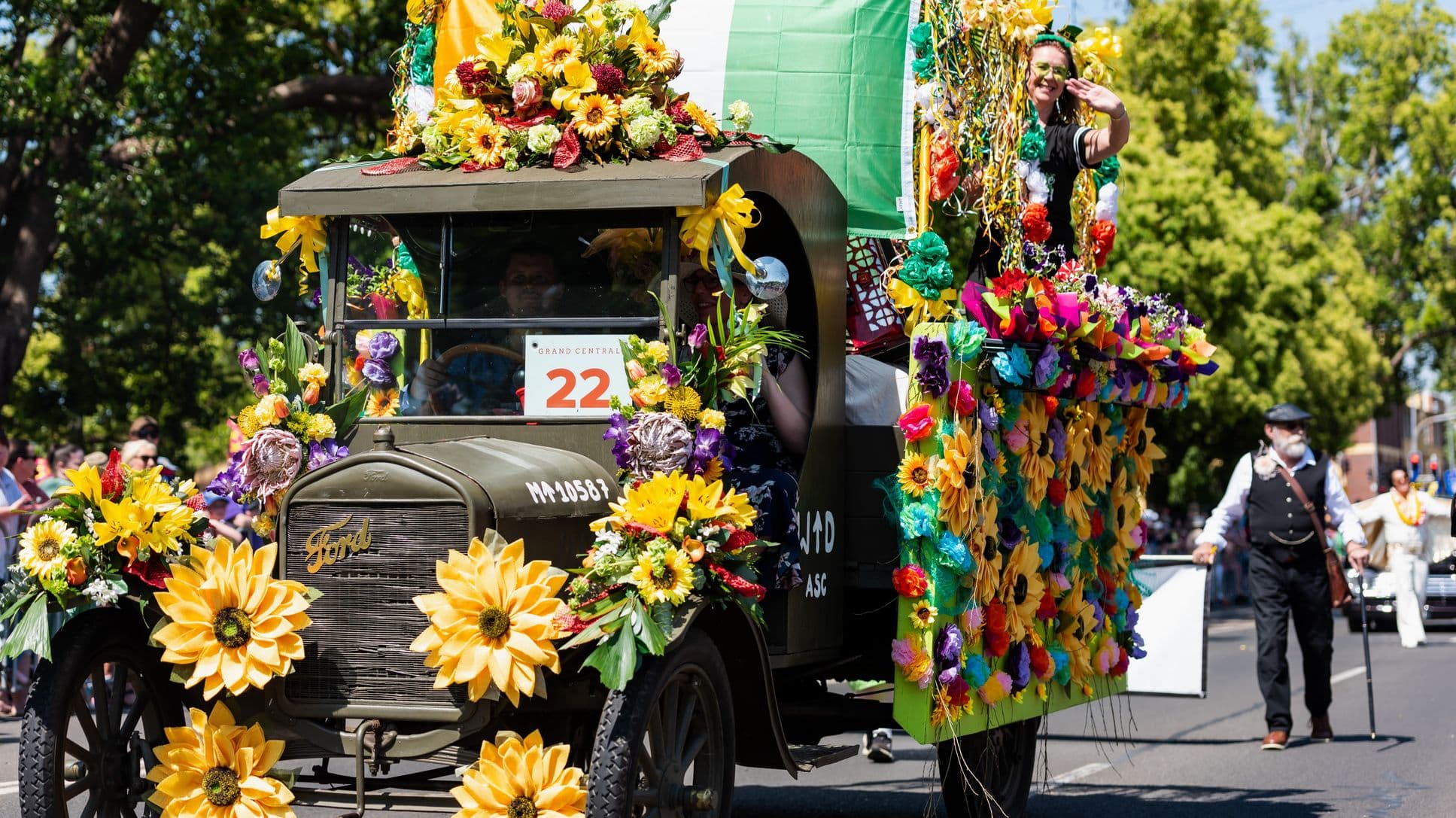 An old Ford vehicle is adorned with vibrant flowers, including large yellow sunflowers, covering the front and sides. A person is seated inside, partially visible. On the right, a woman stands on a colourful float attached to the vehicle, waving. The float is decorated with multicoloured flowers and ribbons. In the background, a man with a cane walks on the road, and trees and spectators line the street.