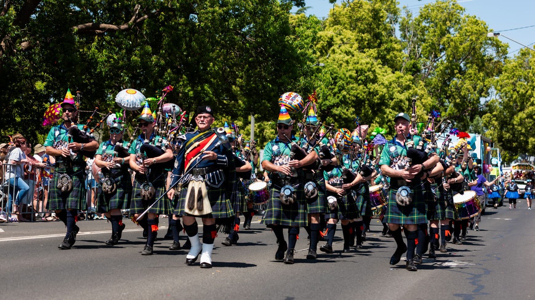 A parade of bagpipers in kilts and colourful party hats marches down a street. They are playing bagpipes and some are carrying drums. The group is dressed in green shirts and tartan kilts. Onlookers stand behind barriers on the left side of the street, watching the procession. Trees with lush green foliage line the background, providing shade and a vibrant backdrop to the scene.