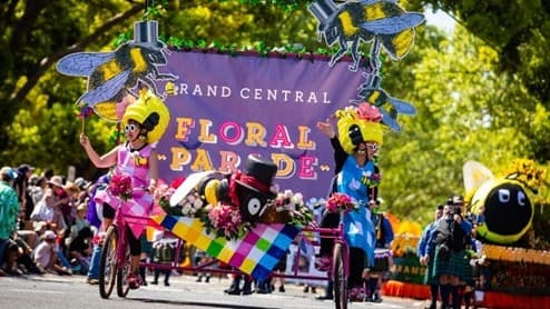A vibrant parade scene with participants in colourful costumes riding bicycles. On the left, a person in a pink outfit with a large headpiece rides a bike. In the centre, a person in a multicoloured outfit pushes a cart adorned with flowers. On the right, another person in a blue costume rides a bike. Behind them, a large purple banner reads "Grand Central Floral Parade" with decorative elements. The background is filled with trees and spectators lining the street.