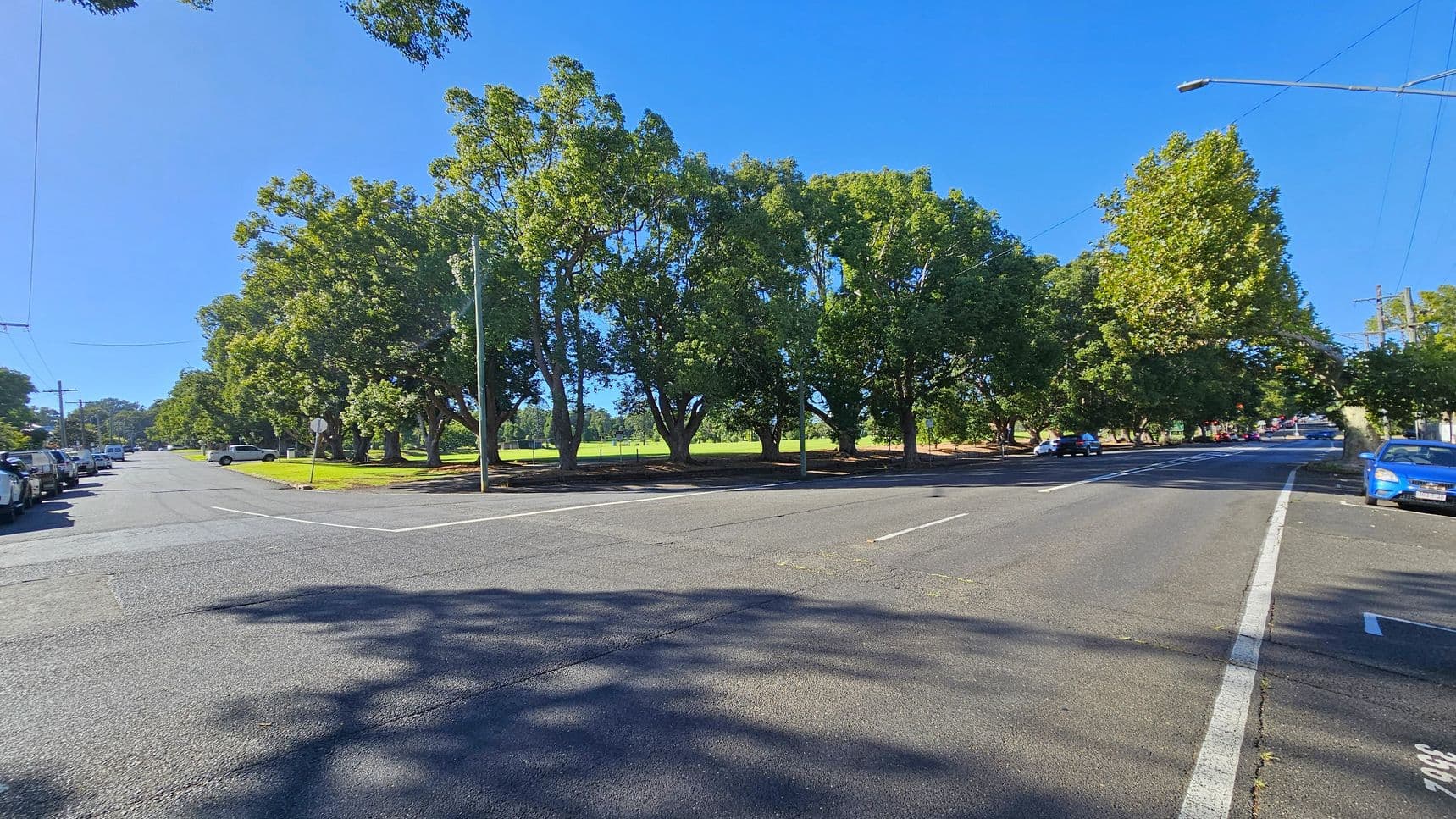 A street intersection with a wide road in the foreground. On the left, cars are parked along the kerb, and a row of large, leafy trees lines the street. On the right, more trees and parked cars are visible. In the background, a grassy area is partially visible through the trees. The road surface is smooth, with shadows from the trees cast across it. A streetlight and traffic sign are present near the centre.