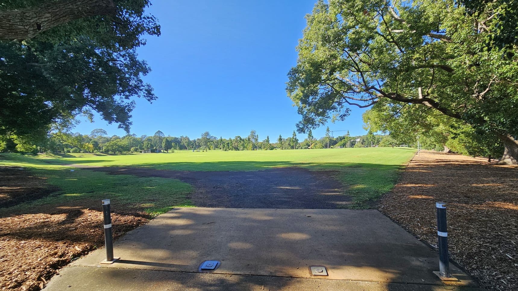 A park scene with a concrete path in the foreground, flanked by two metal posts. The path leads to a grassy field in the middle, surrounded by trees. On the left and right, large trees with dense foliage provide shade. The ground beneath the trees is covered with mulch. In the background, more trees line the horizon, creating a lush, green landscape.