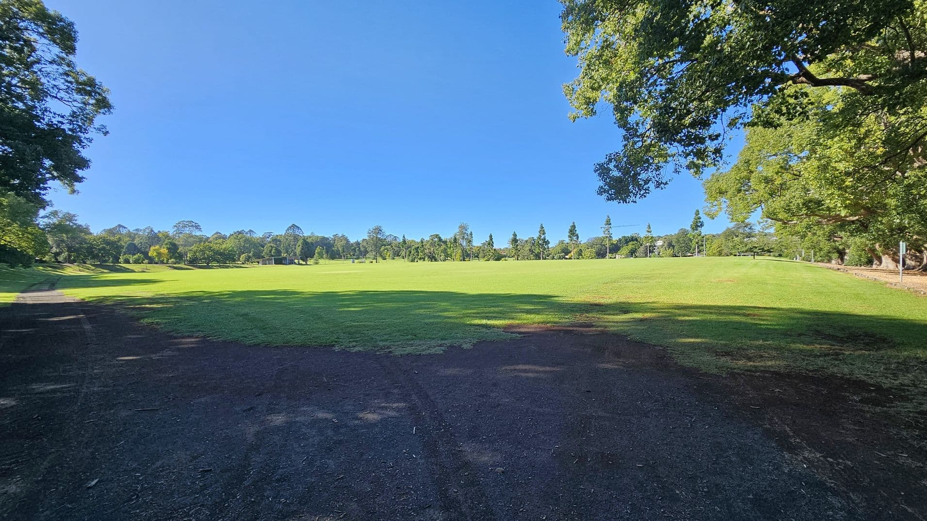A wide, open grassy field stretches across the centre of the image, bordered by a variety of trees in the background. On the left and right, large trees with dense foliage frame the scene, casting shadows on the grass. The foreground features a dark, earthy path leading into the field. The overall setting is a spacious, green parkland.