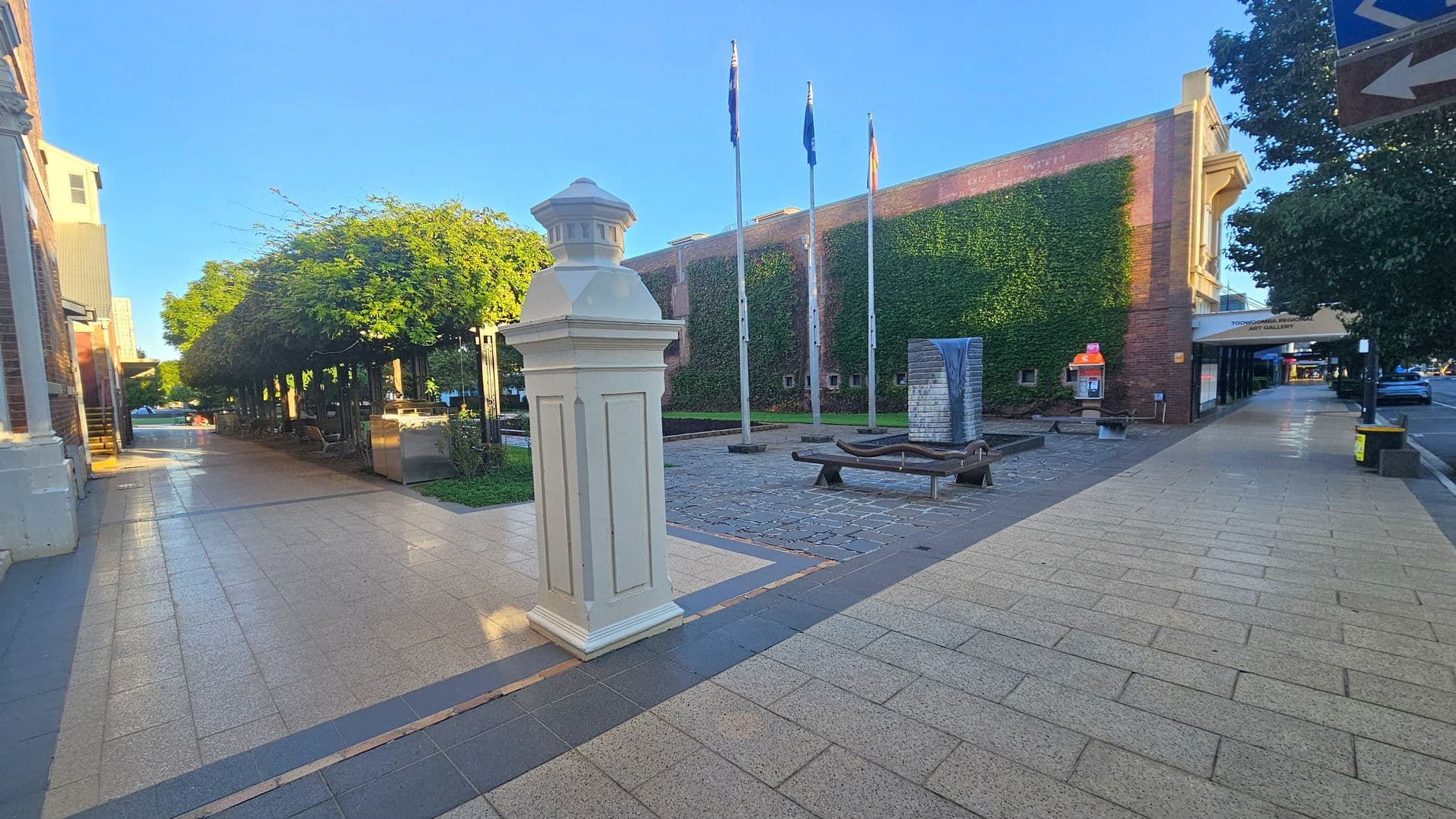 A paved walkway with a white post in the foreground. On the left, a row of trees provides shade over benches. In the centre, three flagpoles stand in front of a brick wall covered in ivy. A modern sculpture with a water feature is nearby. On the right, a building with a sign for an art gallery is visible, along with a public phone booth. The pavement is light-coloured with darker borders, and there are trees lining the street.