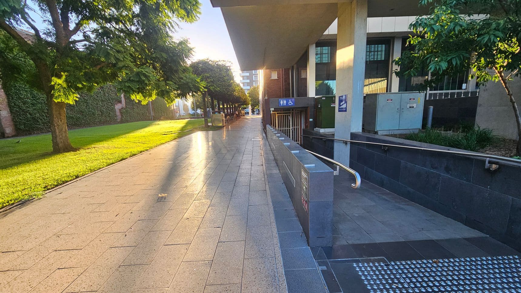 A paved walkway with sunlight casting long shadows. On the left, there's a grassy area with trees lining the path. On the right, a building with a ramp leading to a lower level, marked with signs for toilets and accessibility. The building has large windows and a small garden area with shrubs. In the background, more trees and a distant view of a street with parked cars. The ceiling of the building overhangs the walkway.