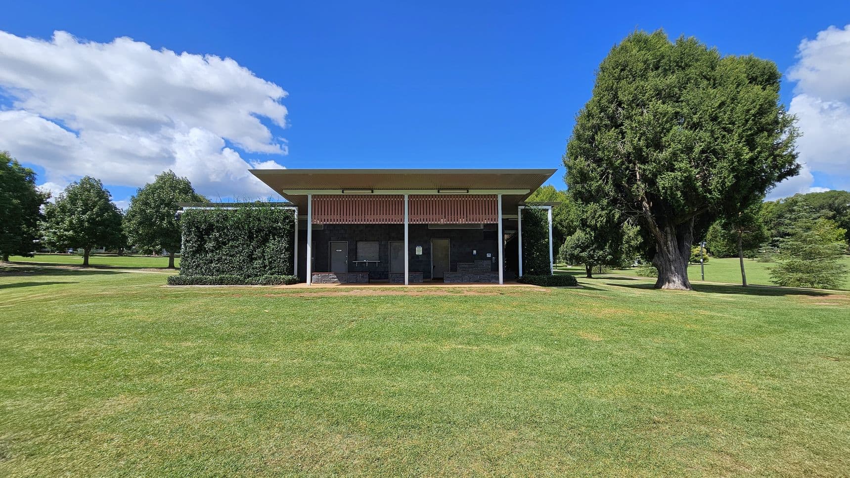 A small, modern building with a flat roof is centred in a grassy park. The structure has a green wall on the left side and several doors and windows on the front. A large tree stands to the right, while smaller trees are scattered in the background. The grass is well-maintained, and the building is surrounded by open space.