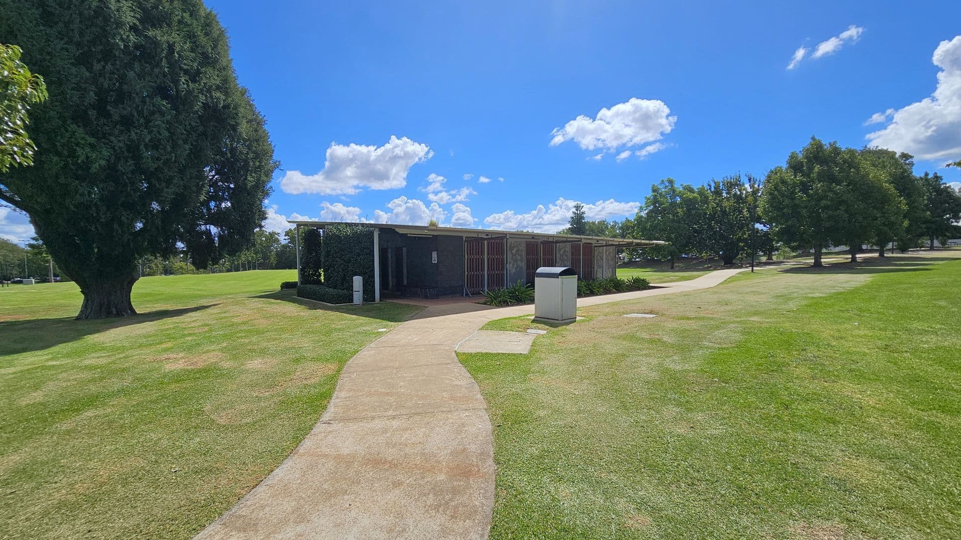 A park scene with a concrete path winding through a grassy area. On the left, a large tree provides shade. In the centre, a low, rectangular building with a flat roof is partially covered with greenery. A bin is positioned near the path. On the right, several trees line the grassy expanse, casting shadows on the ground. The sky is clear with a few clouds.