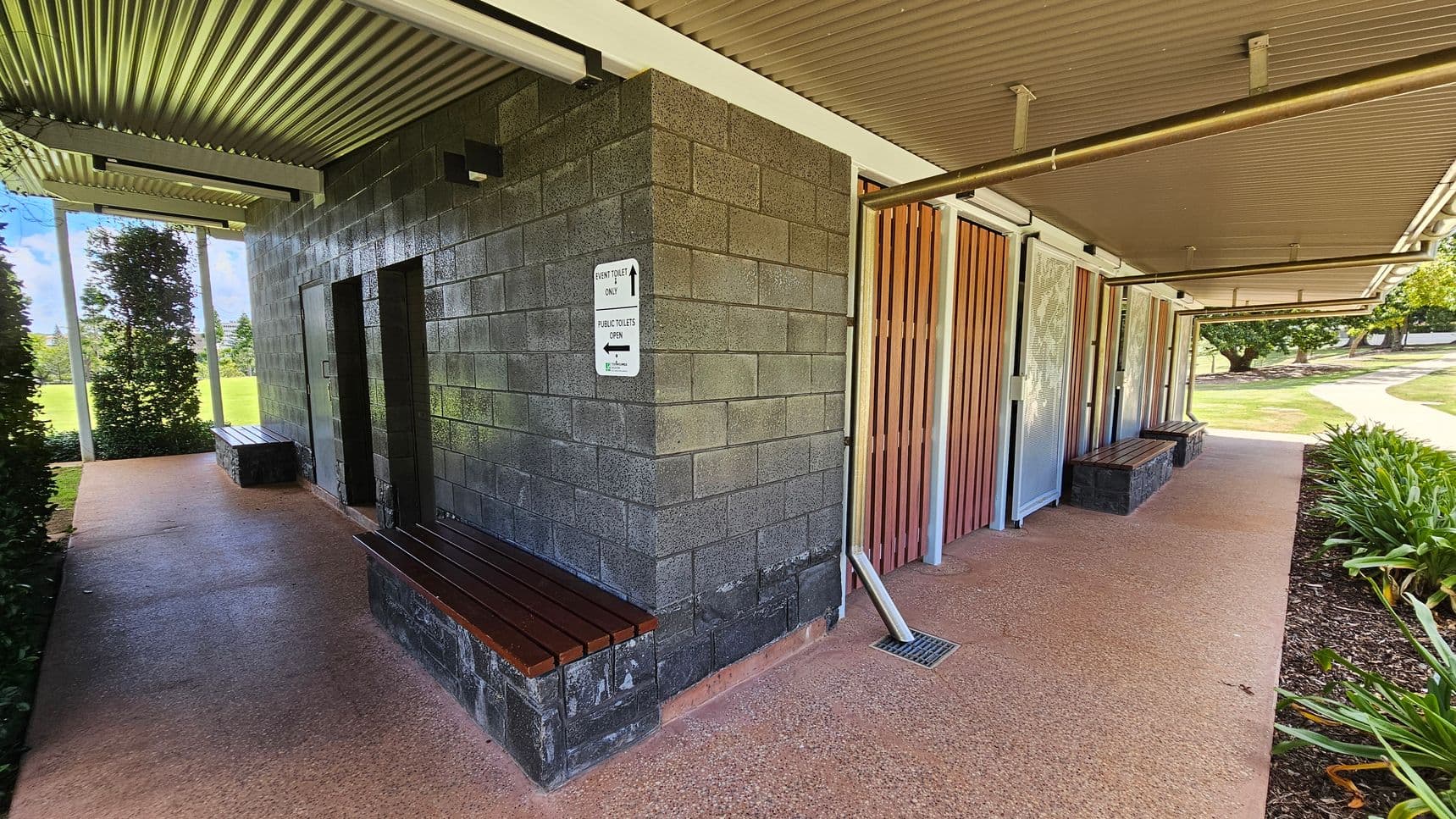 A public toilet facility with a corrugated metal roof. The left side features a dark brick wall with a sign indicating directions for event toilets and public toilets. A wooden bench is attached to the wall. On the right, there are wooden panels and metal doors, with another bench in front. The floor is a textured concrete surface. In the background, there is a grassy area with trees and a pathway, and plants line the right side of the building.