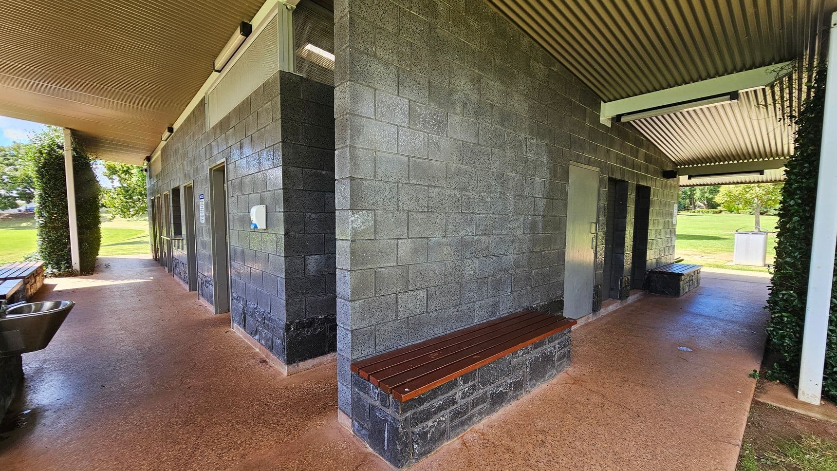 A public restroom facility with grey brick walls and a corrugated metal roof. On the left, there is a stainless steel sink and a wooden bench attached to the wall. The floor is a textured reddish-brown concrete. On the right, there are two doors, likely leading to restrooms, with a soap dispenser mounted on the wall. In the background, there is a grassy area with trees and a bin. The ceiling is lined with metal panels.
