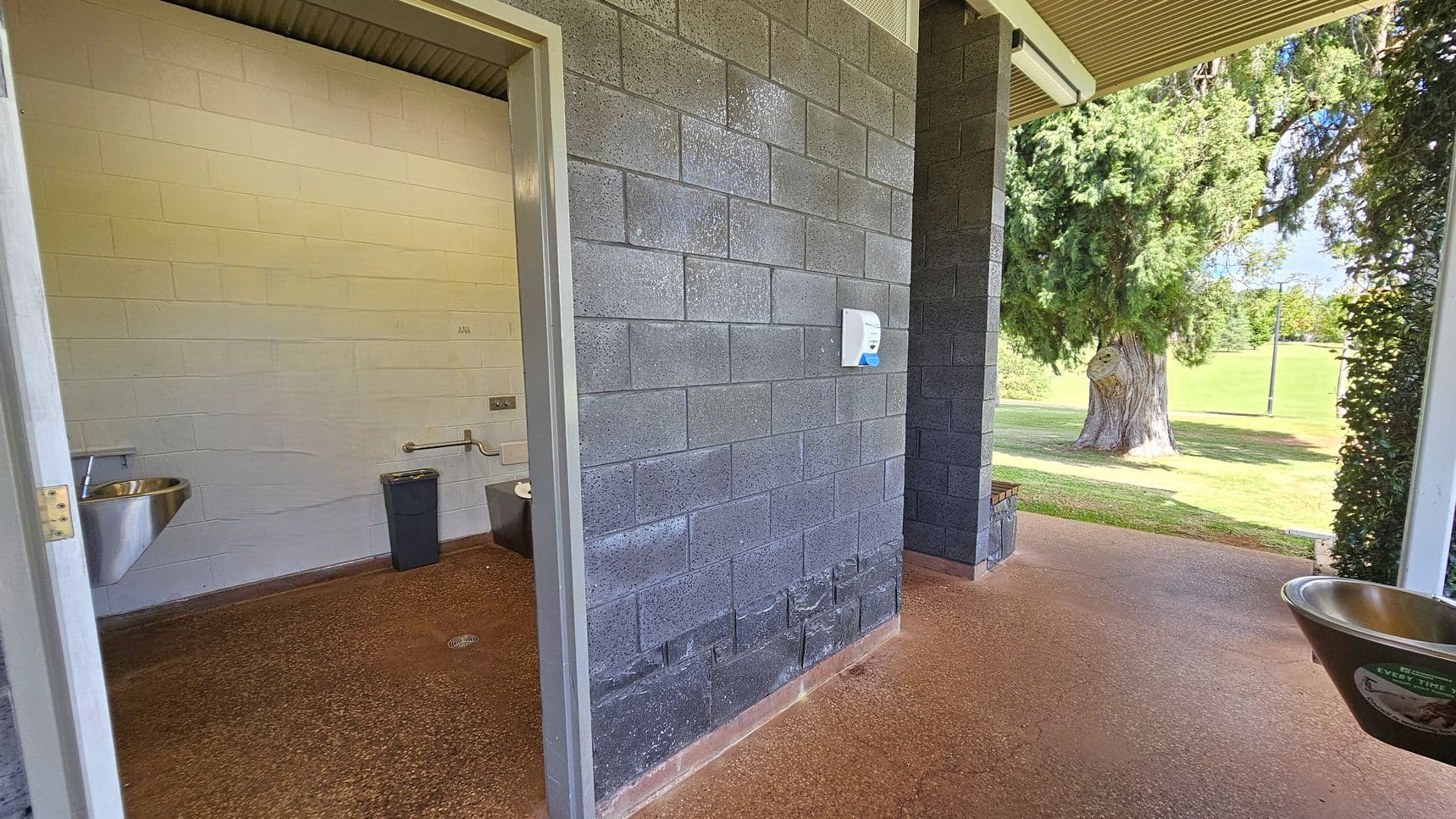 A public restroom with a grey brick exterior wall and an open door leading to the interior. Inside, there is a stainless steel sink and a waste bin. The floor is brown and textured. Outside, a large tree and grassy area are visible. A soap dispenser is mounted on the exterior wall. The ceiling is corrugated metal.