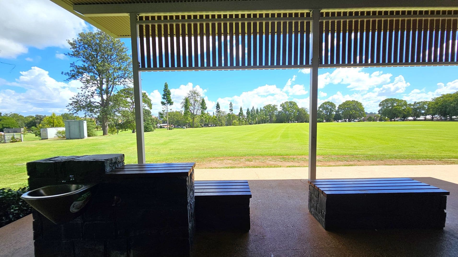 A covered outdoor area with a view of a large grassy field. In the foreground, there are two benches and a sink on the left. The ceiling has a slatted design, allowing light to filter through. In the background, the field is bordered by trees, and there are a few structures visible to the left. The ground is paved with a concrete path running parallel to the field.