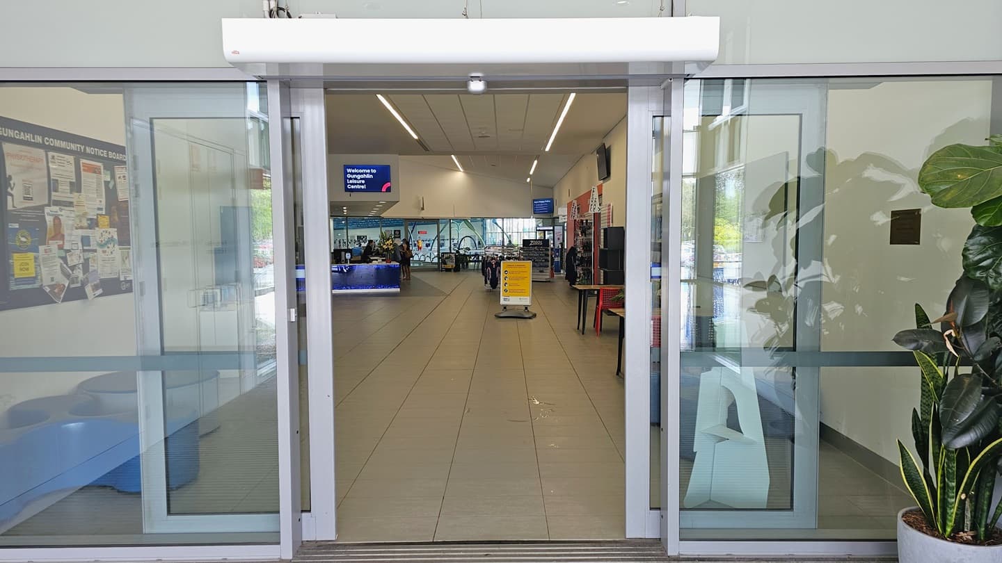 The entrance comprises automatic glass doors. Pots of snake plants and ornamental plants are positioned on the right. Inside, a free-standing banner is placed in the middle. To the left, cushioned seating and a board filled with flyers are present. To the right, movable plastic tables and chairs are arranged. Further in is the reception desk with a flatscreen TV displaying a welcome message.