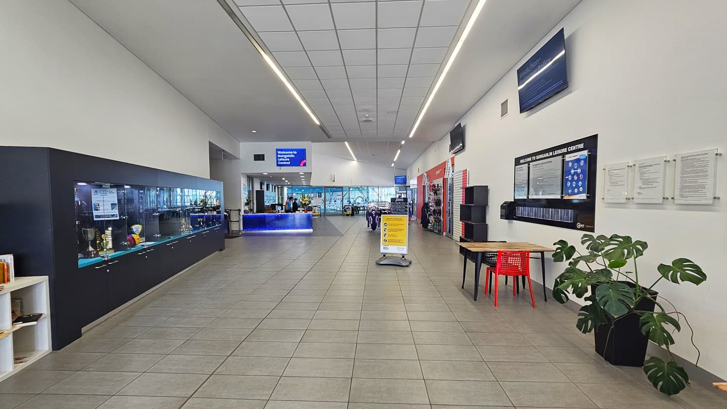 On the left is a spacious, long open corridor with a large glass display rack showcasing the venue's achievements—trophies, awards, and a collection of books. To the right is a pot of monstera plant. The right wall displays facilities information, adjacent to a movable table and chairs. A flat-screen TV is mounted on top. Further in is the reception desk, with a free-standing banner positioned near the wooden display rack.