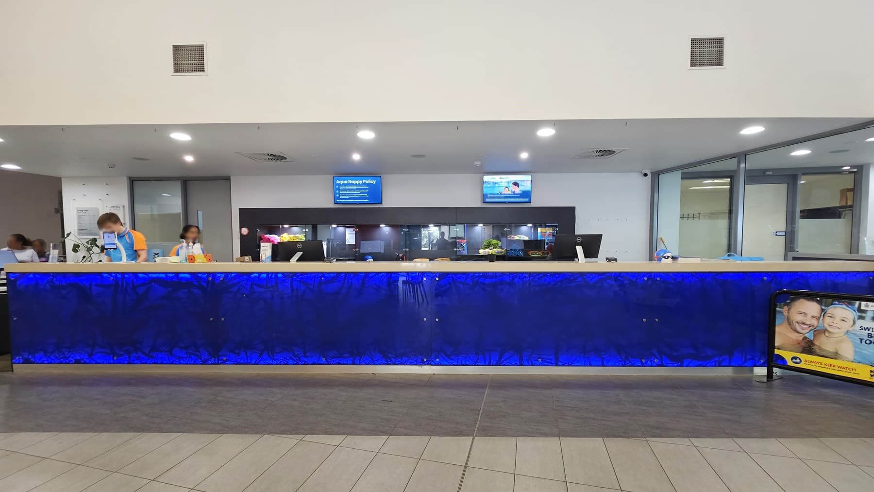In the middle is the reception desk, featuring distinctive royal blue panels with staff behind the counter. A display rack is positioned behind the counter. Two flat screen TVs are mounted on the wall. A banner is positioned on the right. The ceiling is all white with fluorescent lights, and the floor has grey tiles.