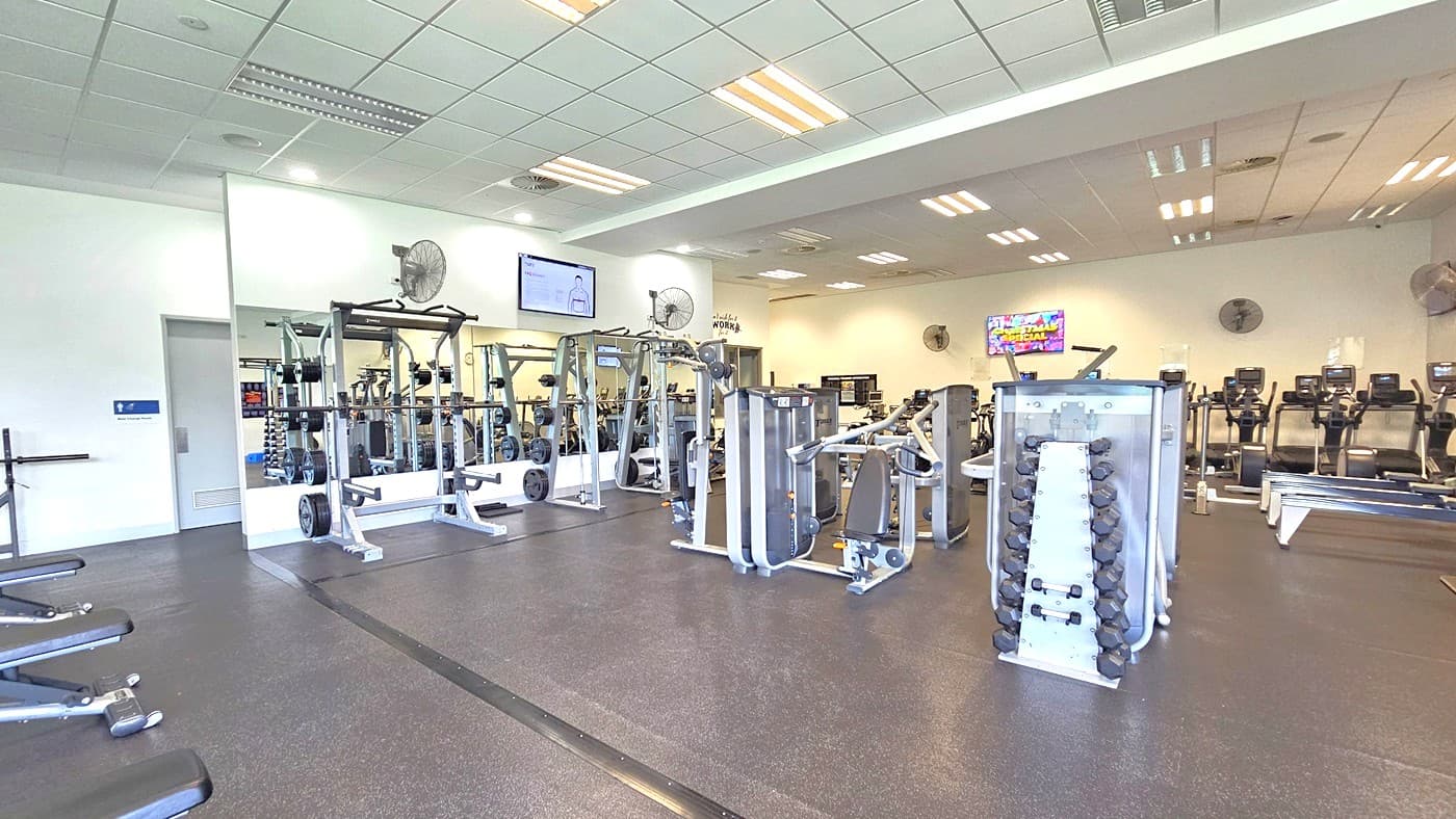 On the left, are bench press machines; in the middle, Smith Machines by a mirror, surrounded by free weights and resistance machines. On the right are row machines and cardio machines. An accessible bathroom is located on the left, behind the middle wall. Black rubber tiles cover the floor. Flat-screen TVs and wall fans are mounted on the upper wall. The ceiling is uniformly white, adorned with fluorescent lights.